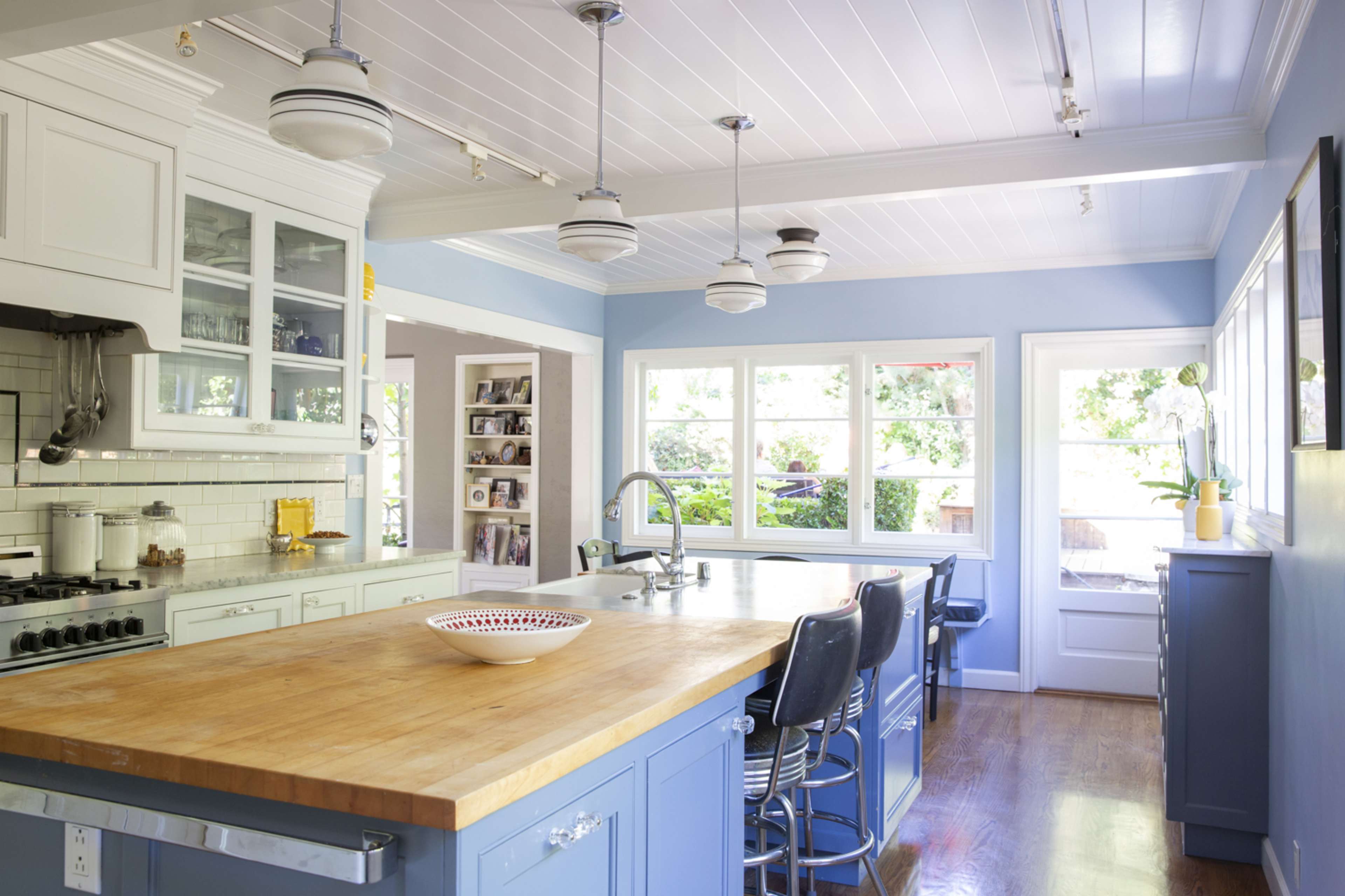A modern kitchen features a large wooden island, blue cabinetry, and multiple pendant lights, with large windows providing natural light.