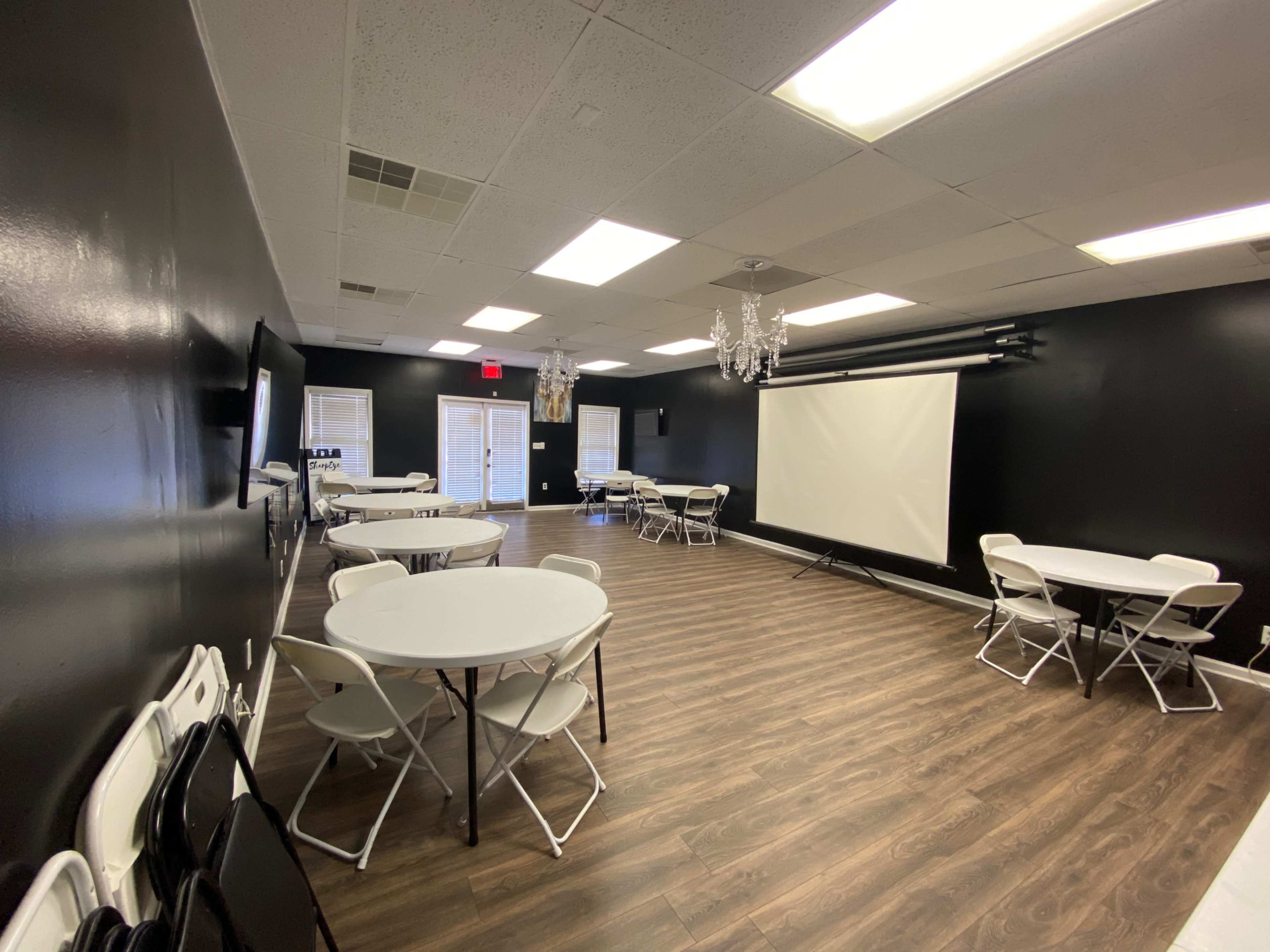 A spacious room with black walls, several round tables with chairs, a large projector screen, and a chandelier.
