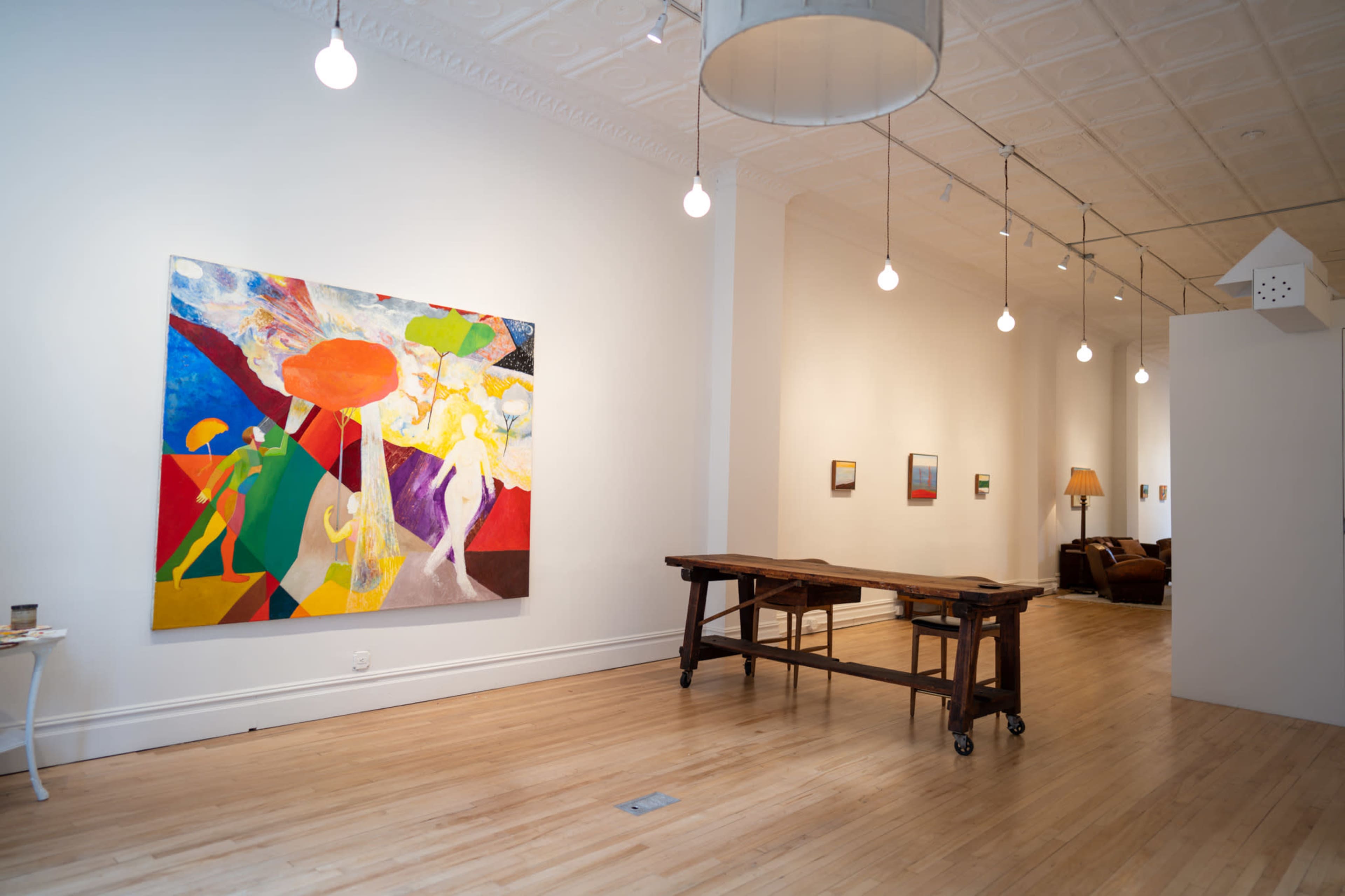 An art gallery with a colorful abstract painting on one wall and a wooden table in the foreground, along with minimalistic lighting and additional artwork displayed in the background.