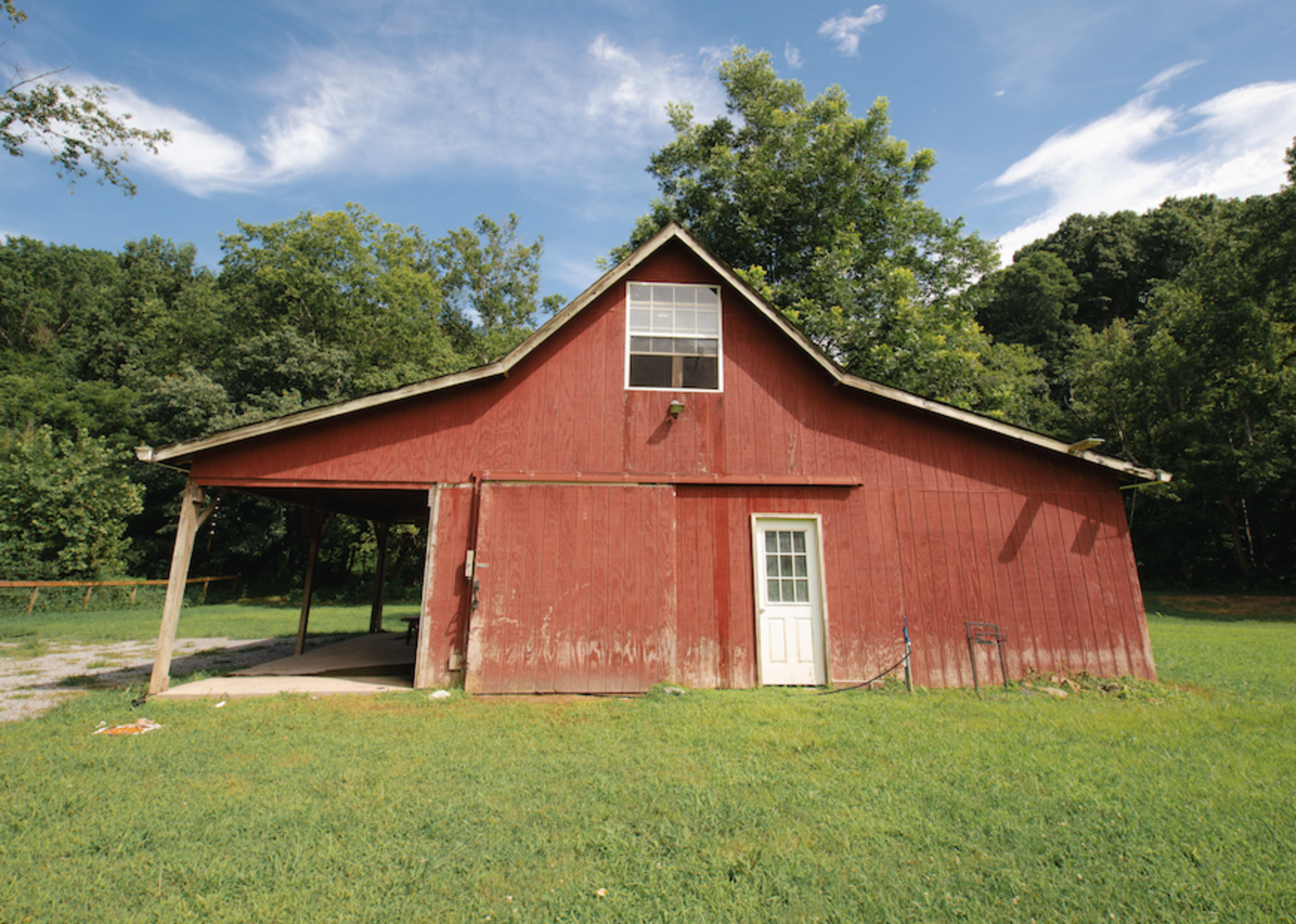 A red barn with a gabled roof sits in a grassy area surrounded by trees under a blue sky.