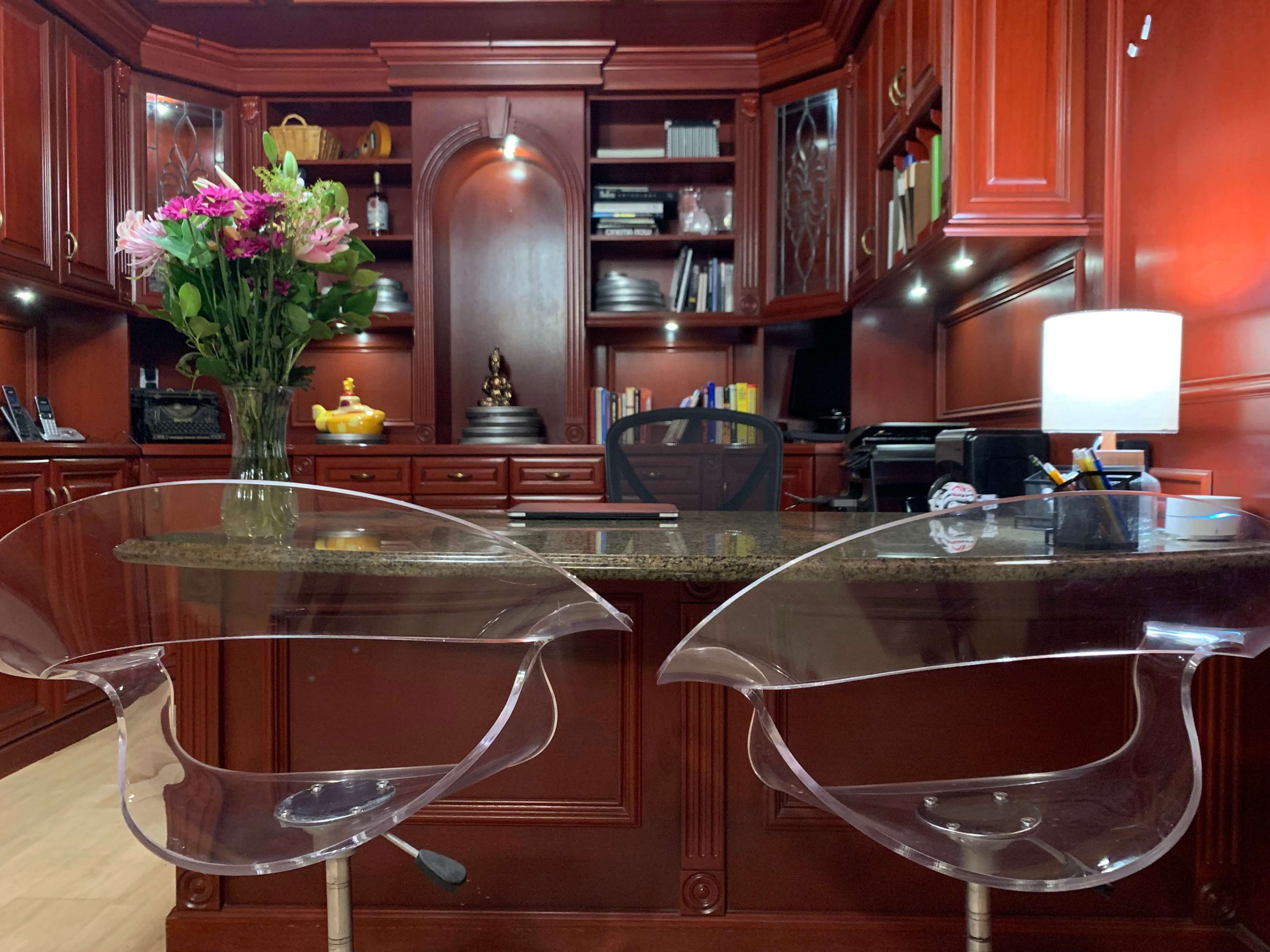 A modern office features a wooden desk with two clear acrylic stools, a floral arrangement, and shelves filled with books and decorative items.