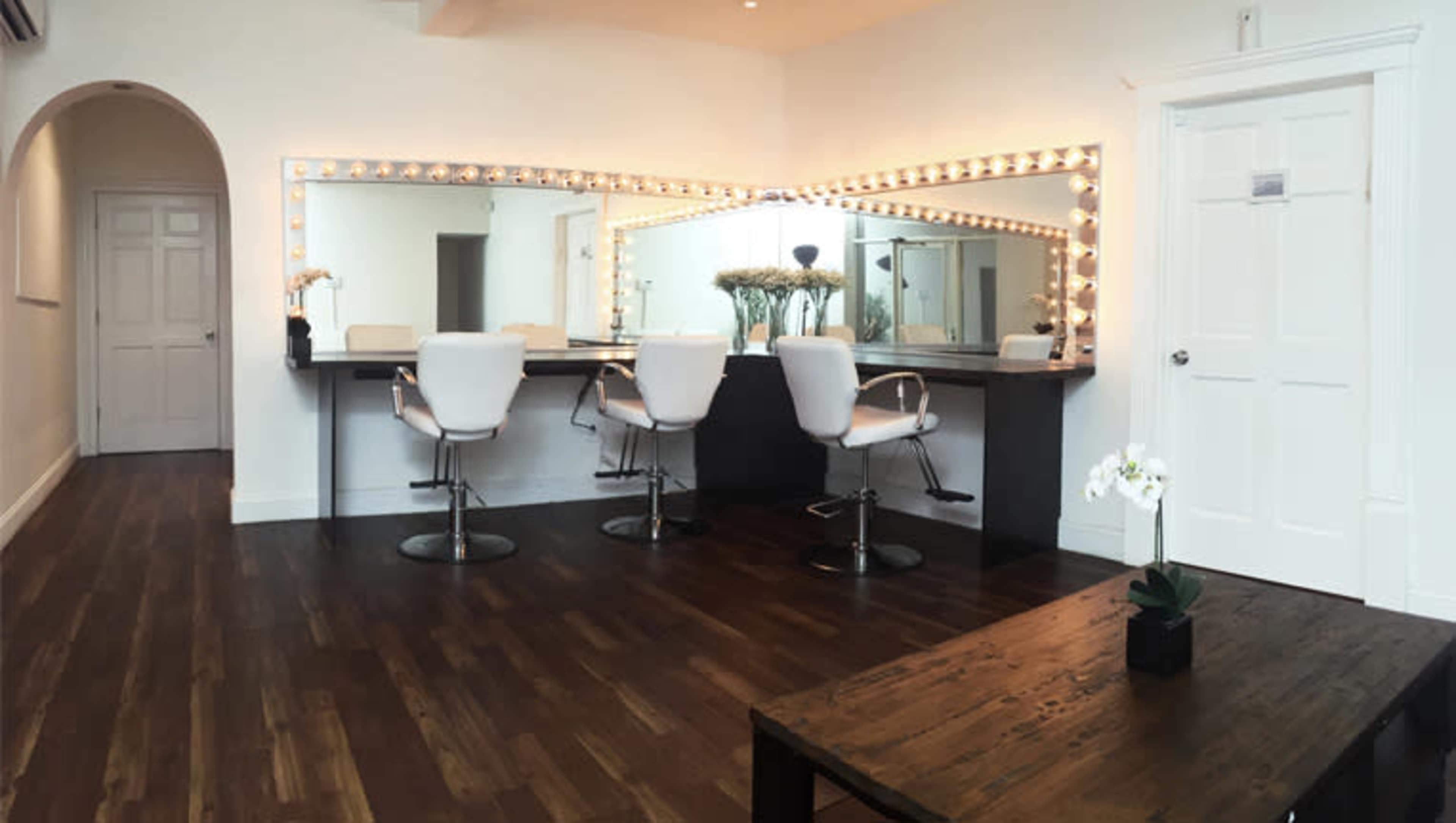 A modern salon interior with a long illuminated mirror, white chairs, and a wooden table on a polished floor.