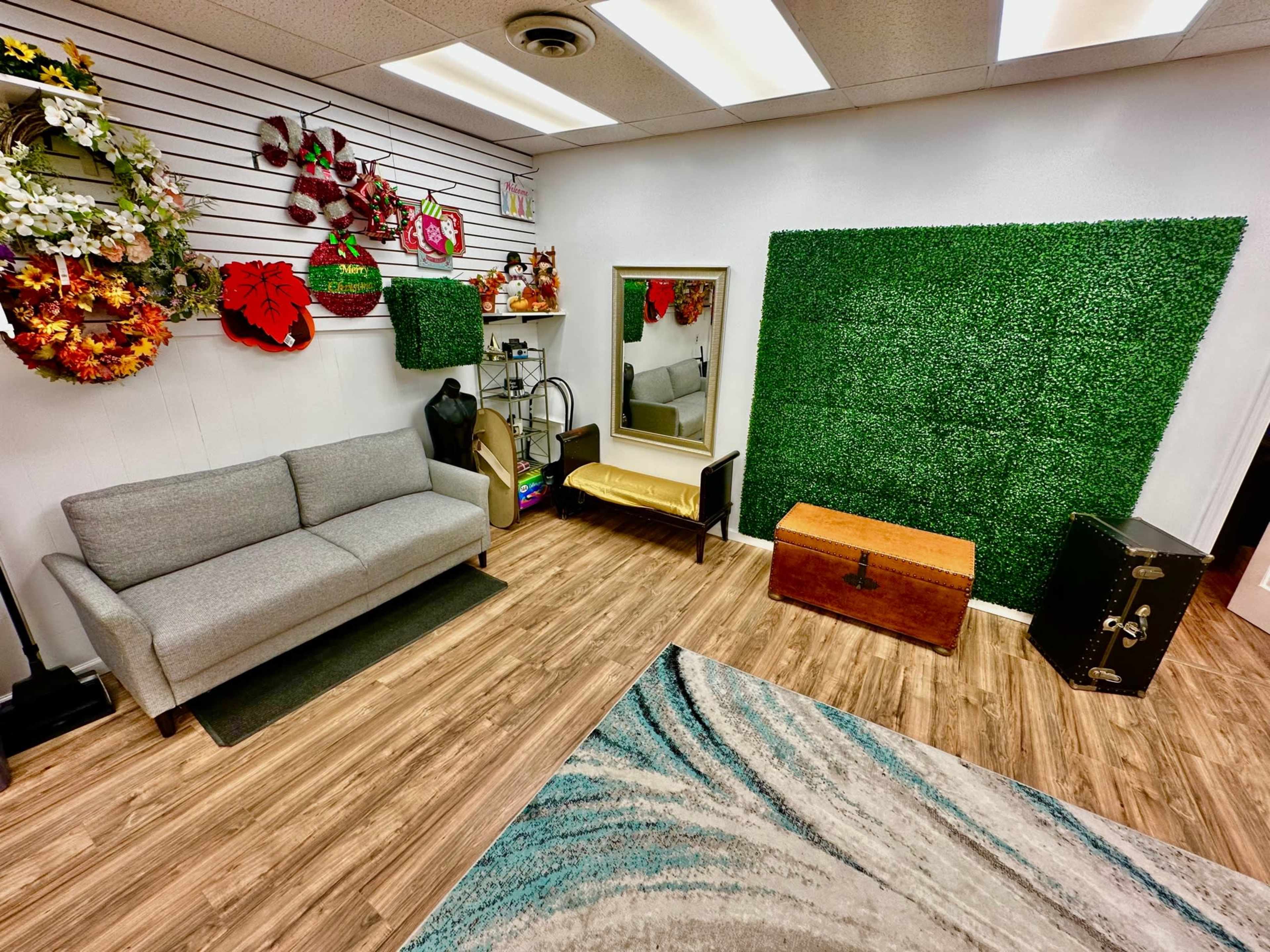 The image shows a decorated interior space featuring a gray sofa, a wooden trunk, a mirror, and a green plant wall, with various floral arrangements on the walls.
