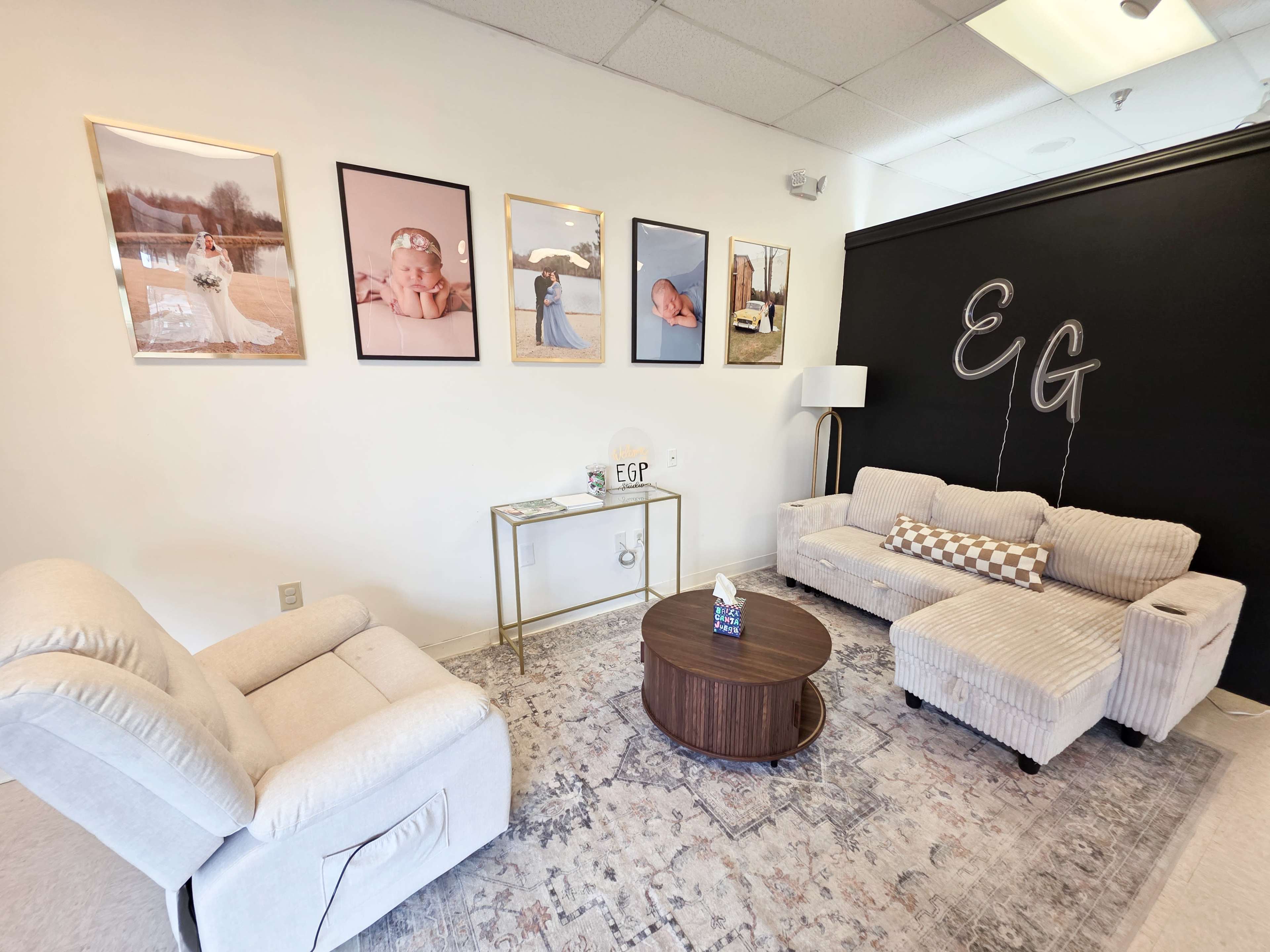 The image shows a cozy room with two seating areas, a round table, and framed photographs on the wall, featuring a neutral color palette and a modern aesthetic.