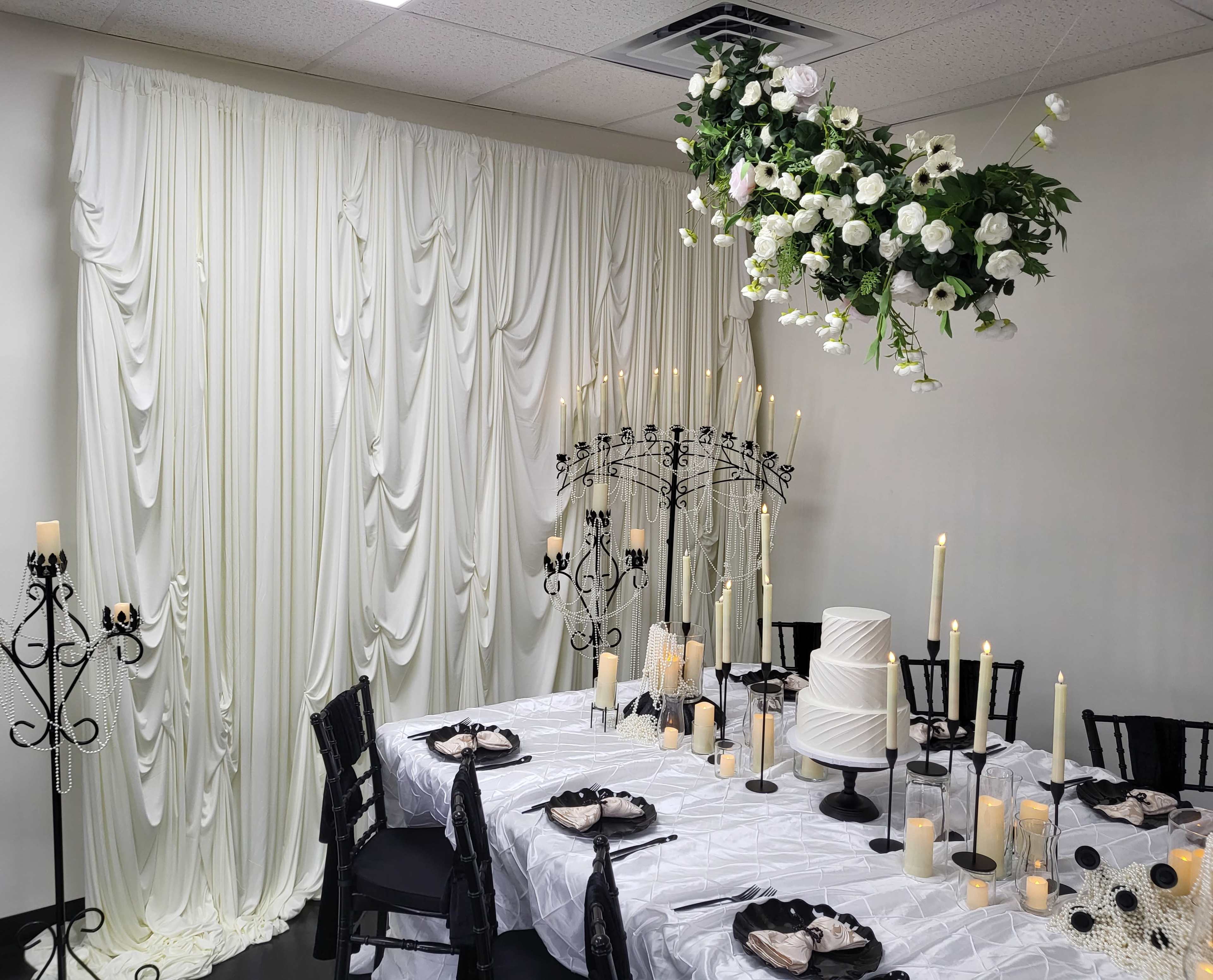 The image depicts an elegantly arranged dining area featuring a white tablecloth, ornate black chairs, floral decorations hanging from the ceiling, and candles set on the table and around the space.