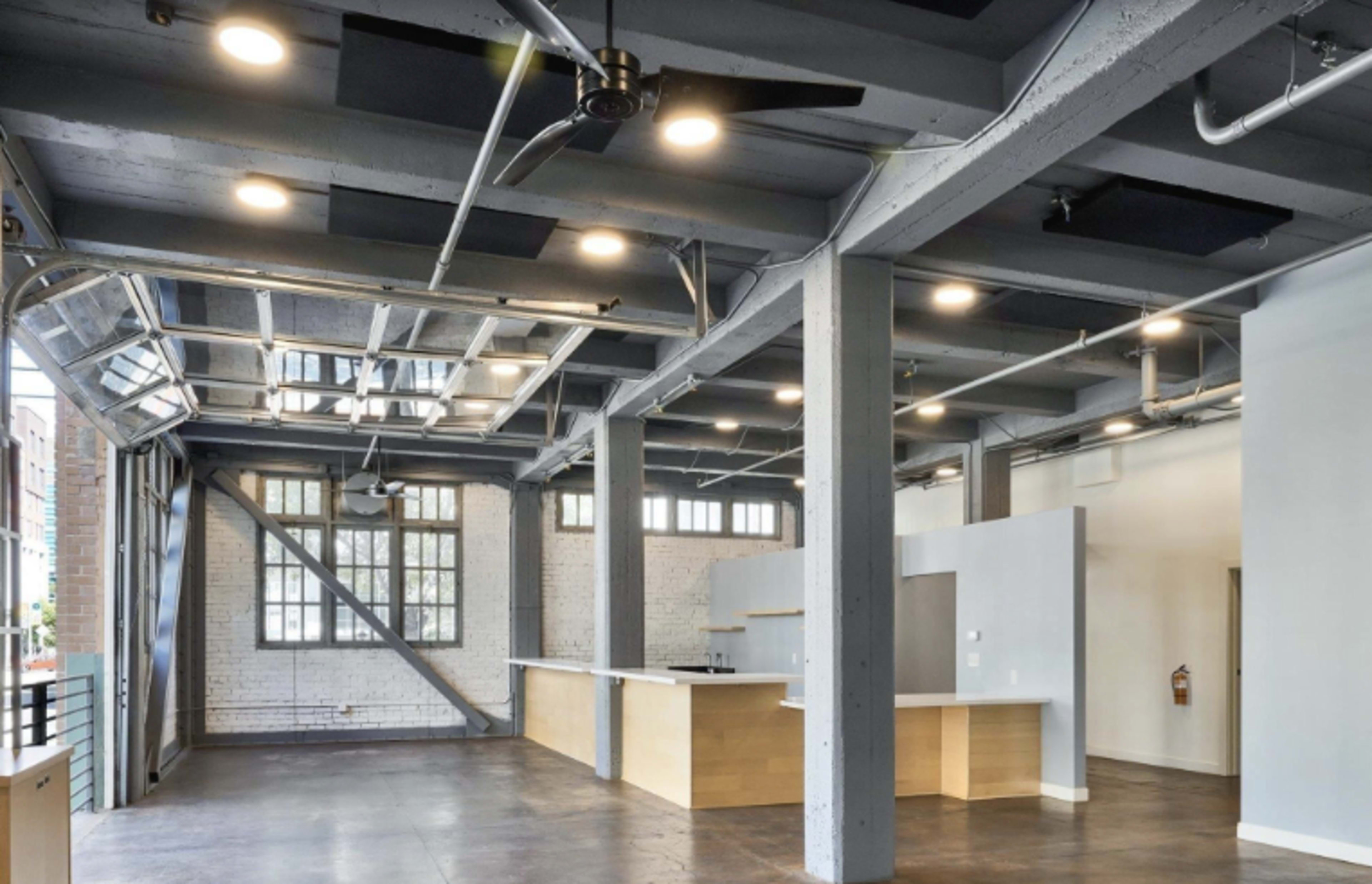 The image shows a spacious, open-concept interior with concrete walls, high ceilings, and large windows, featuring a minimalist kitchen area.