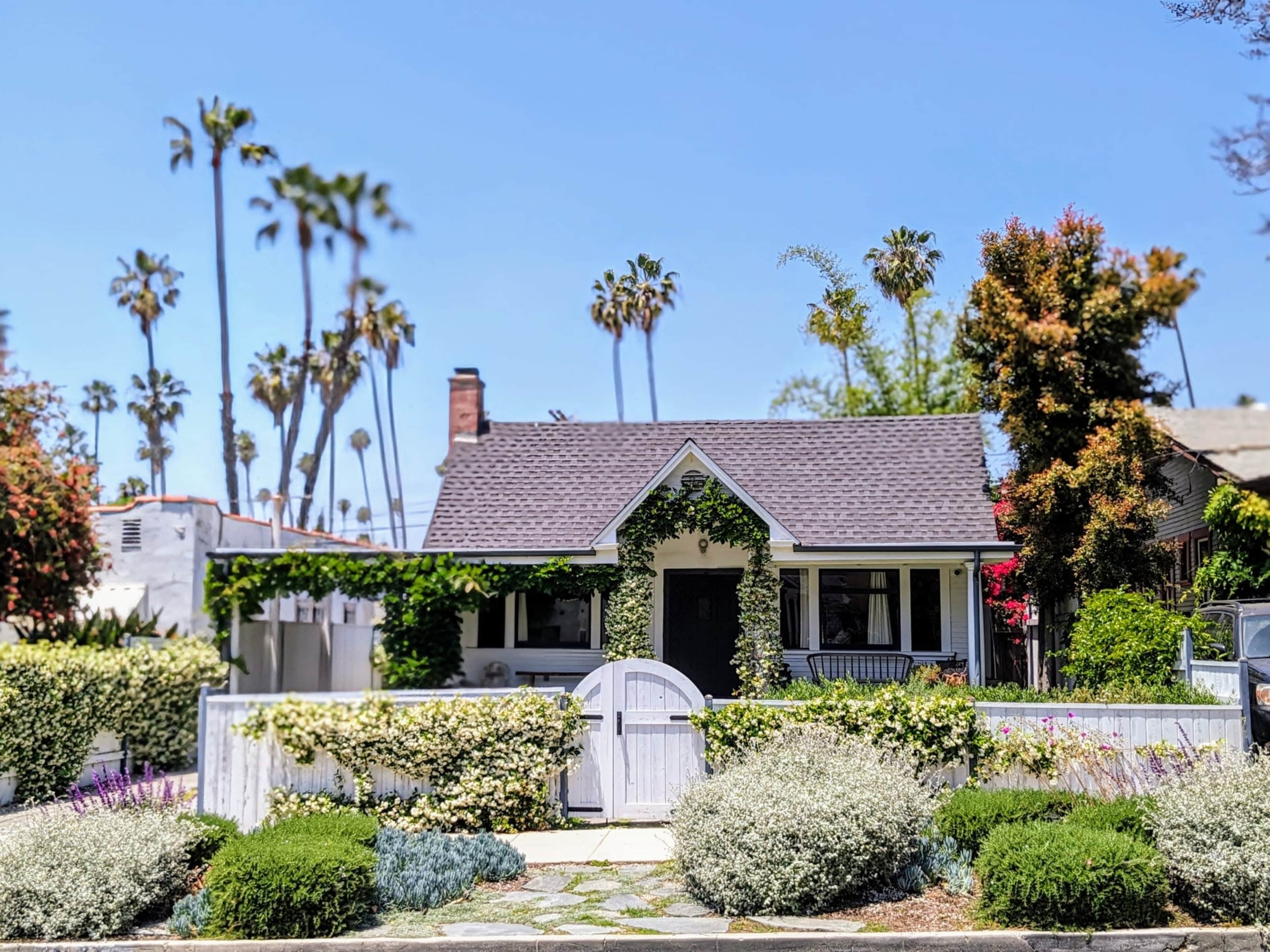 A small white house with a brown roof is surrounded by greenery and blooming flowers, with tall palm trees visible in the background.