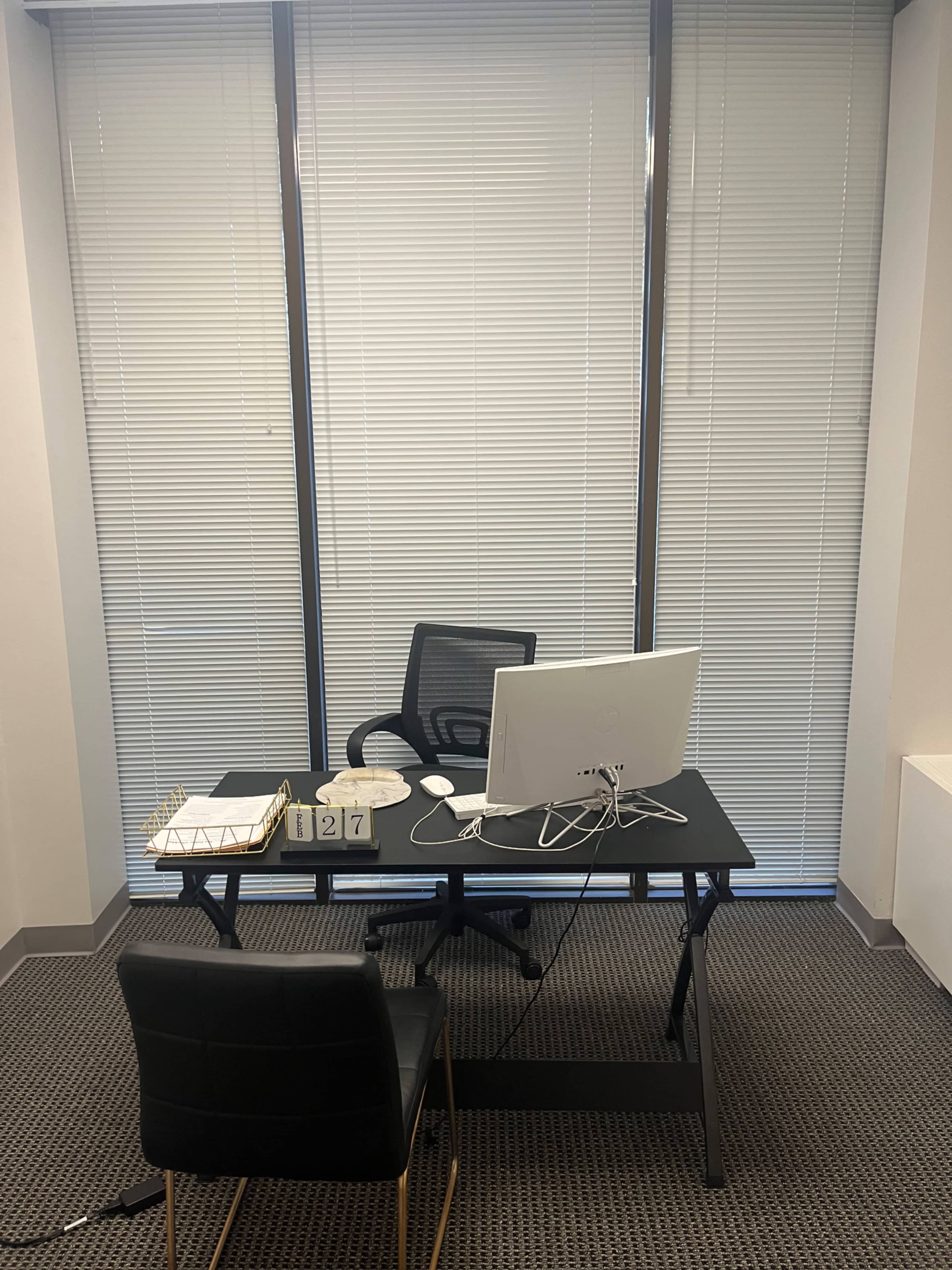 A minimalist office space features a black desk with a computer monitor and an ergonomic chair next to it, set against large windows covered with blinds.