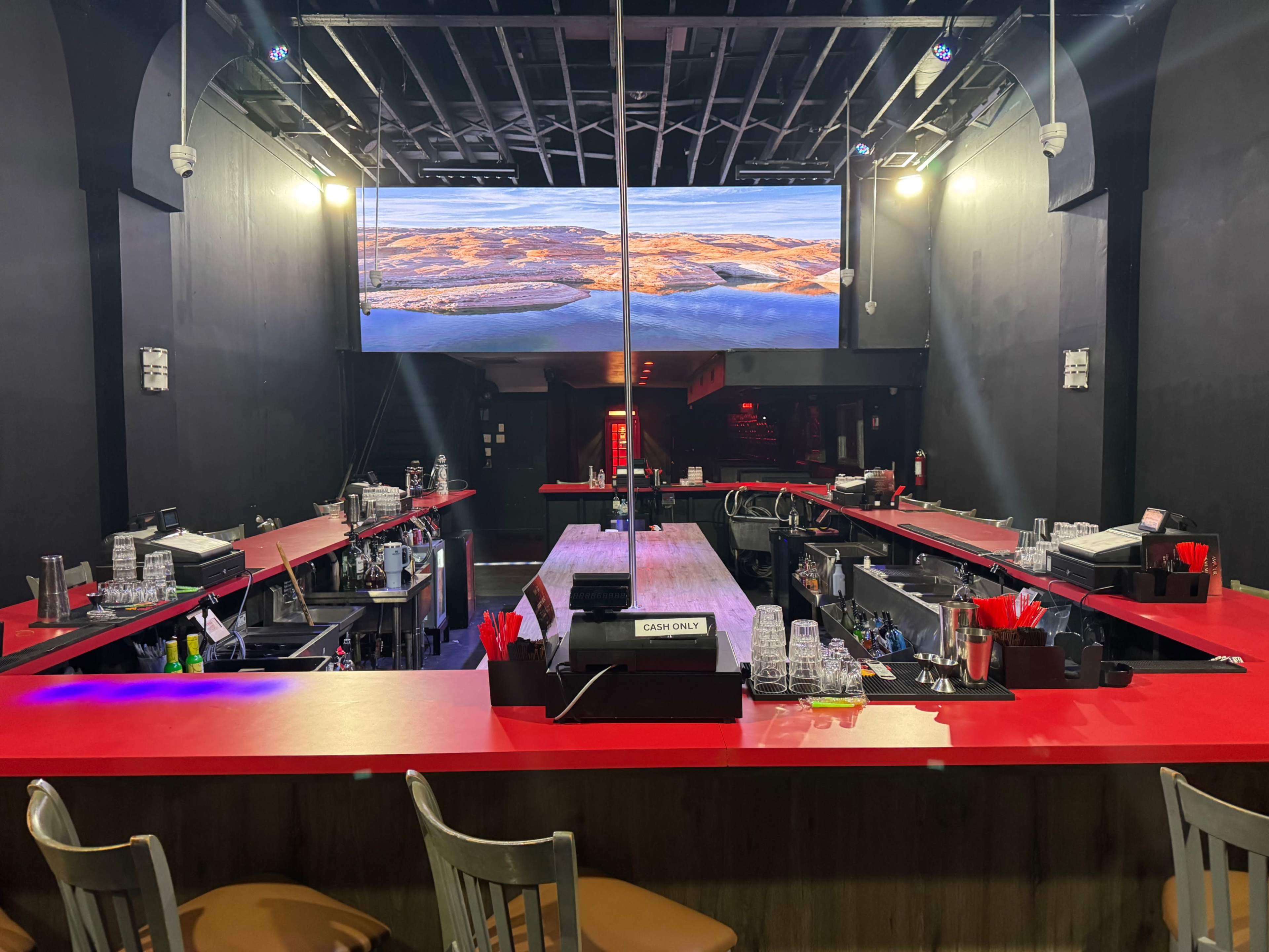 The image shows a modern bar interior with red countertops, multiple bar stations, and a large screen displaying a scenic view.