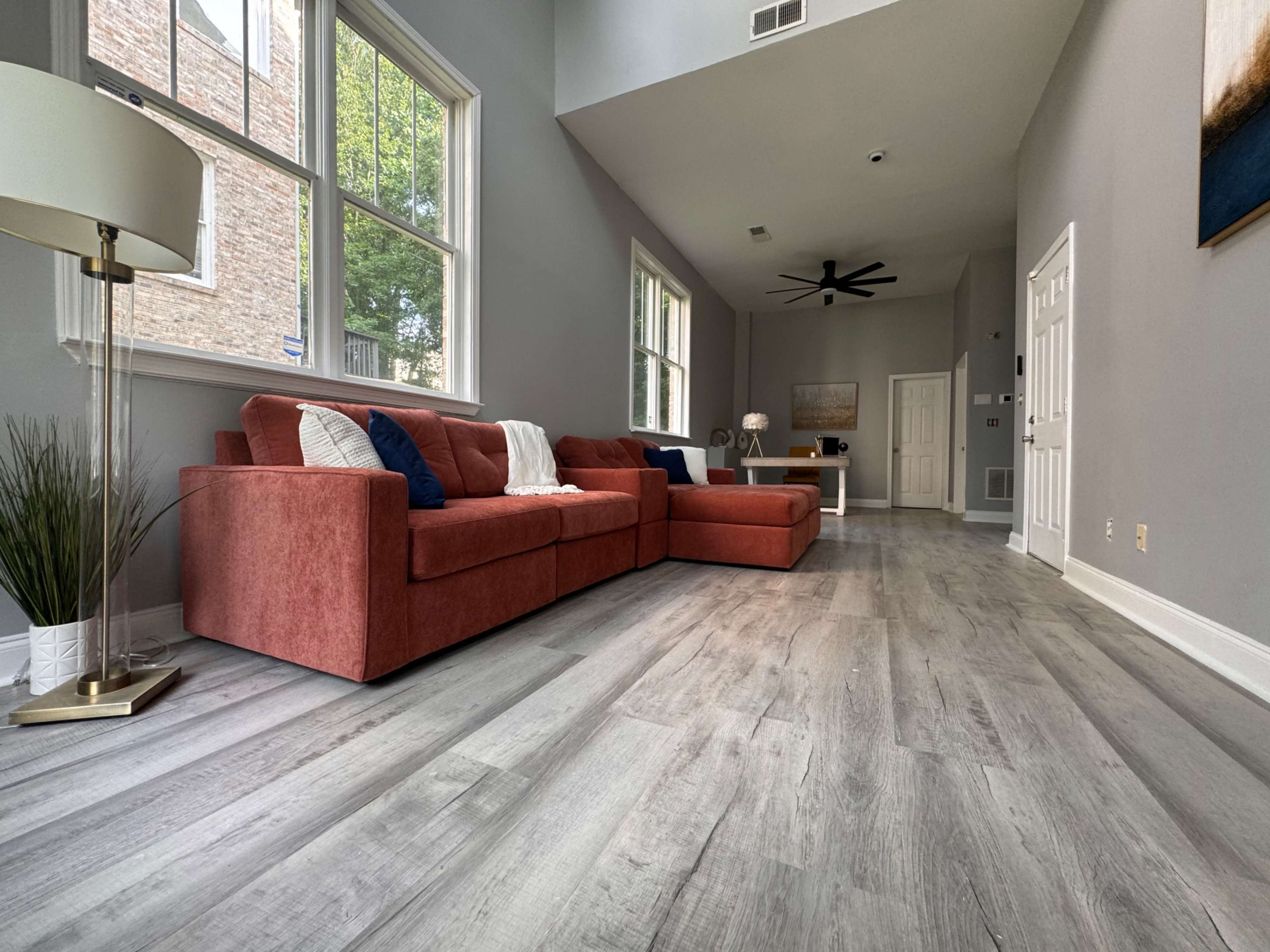 The image shows a spacious living room with a large red sectional sofa, light gray walls, and wooden flooring, featuring large windows that allow natural light to enter.