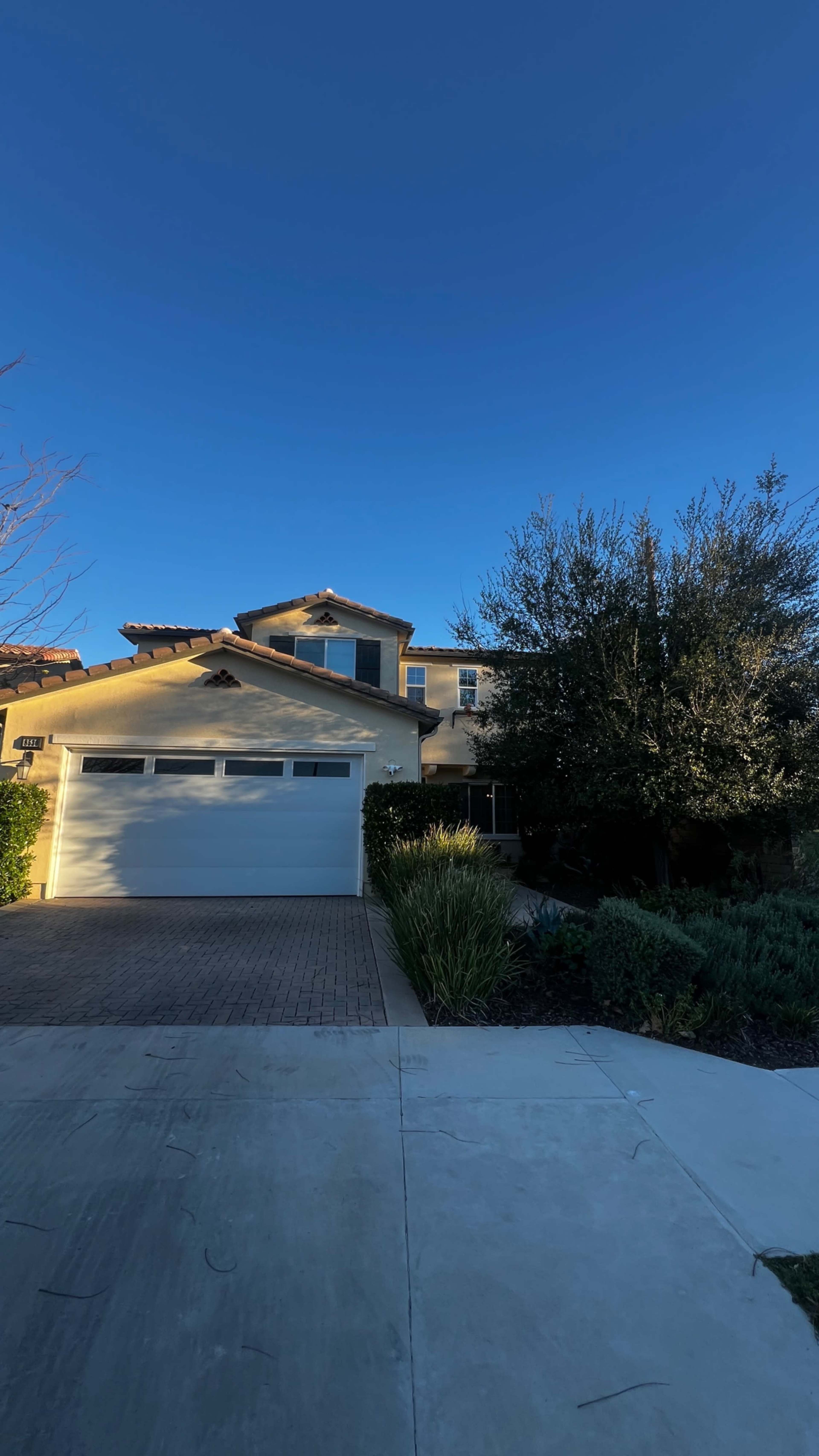 A two-story house with a white garage door and landscaped front yard is illuminated by the afternoon sun against a clear blue sky.