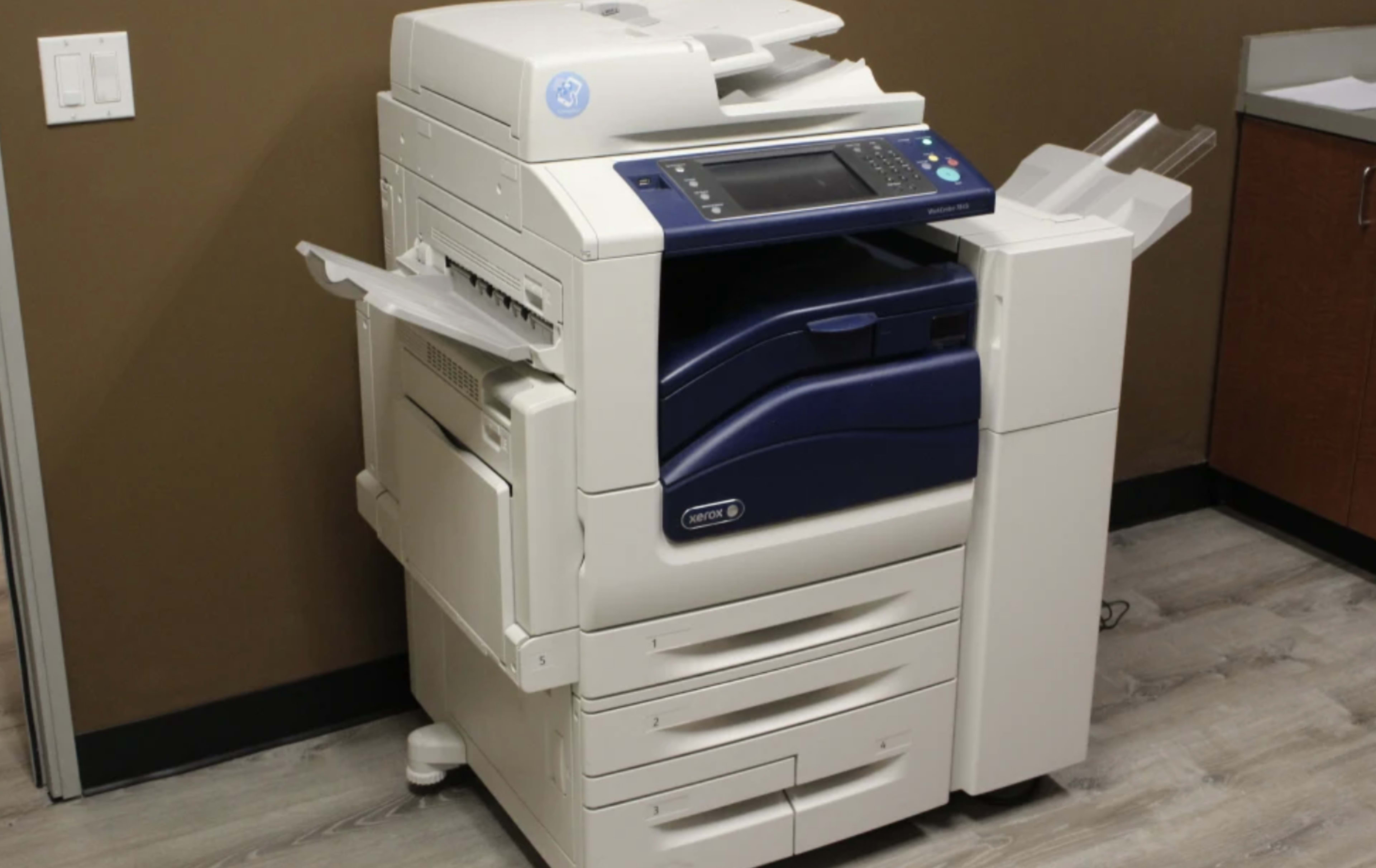 The image shows a multifunction printer with a dark blue front and multiple paper trays, positioned against a brown wall in an office setting.