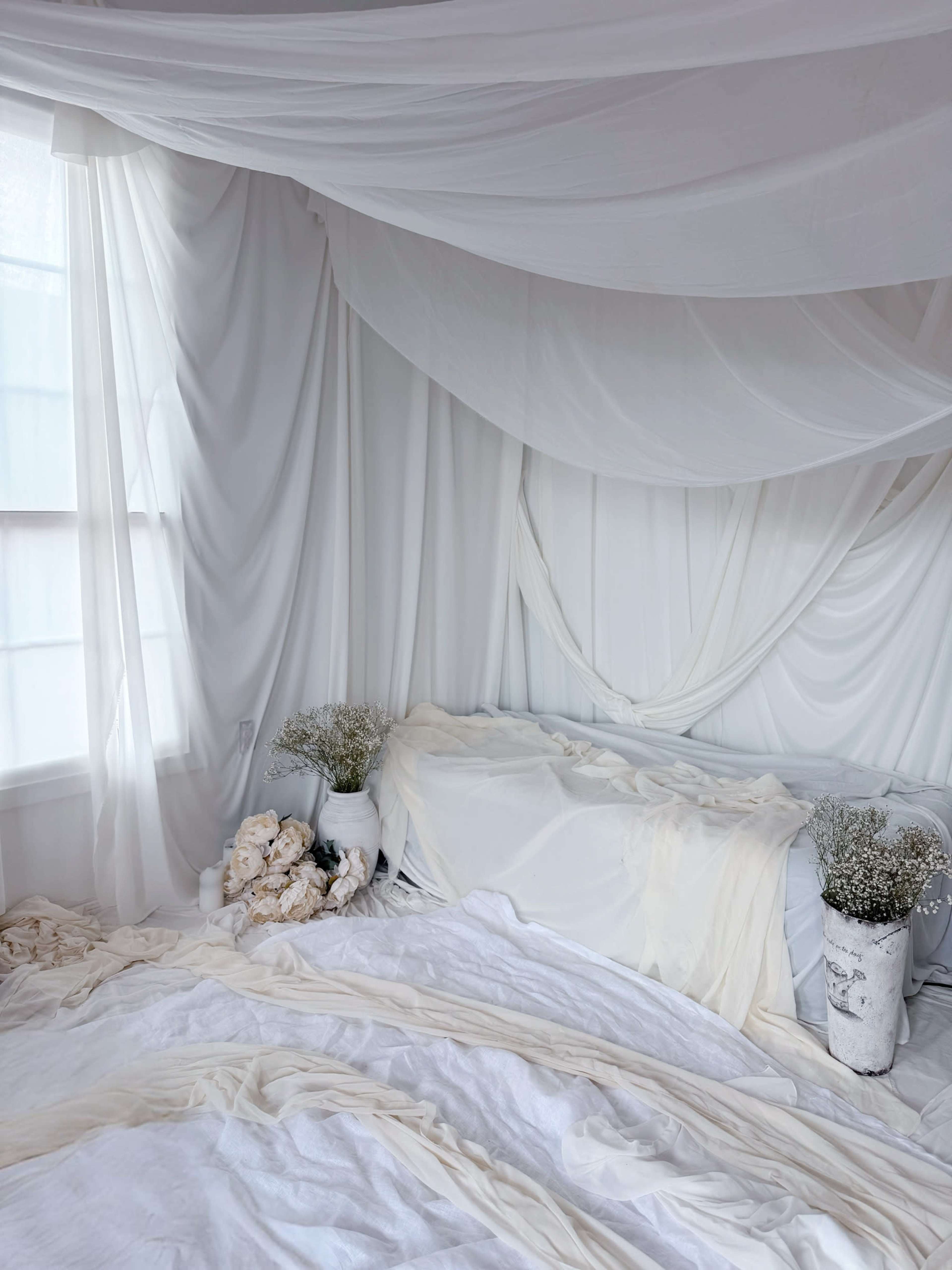 The scene features a bedroom with white draped fabric, a bed covered in white linens, and two vases filled with flowers.