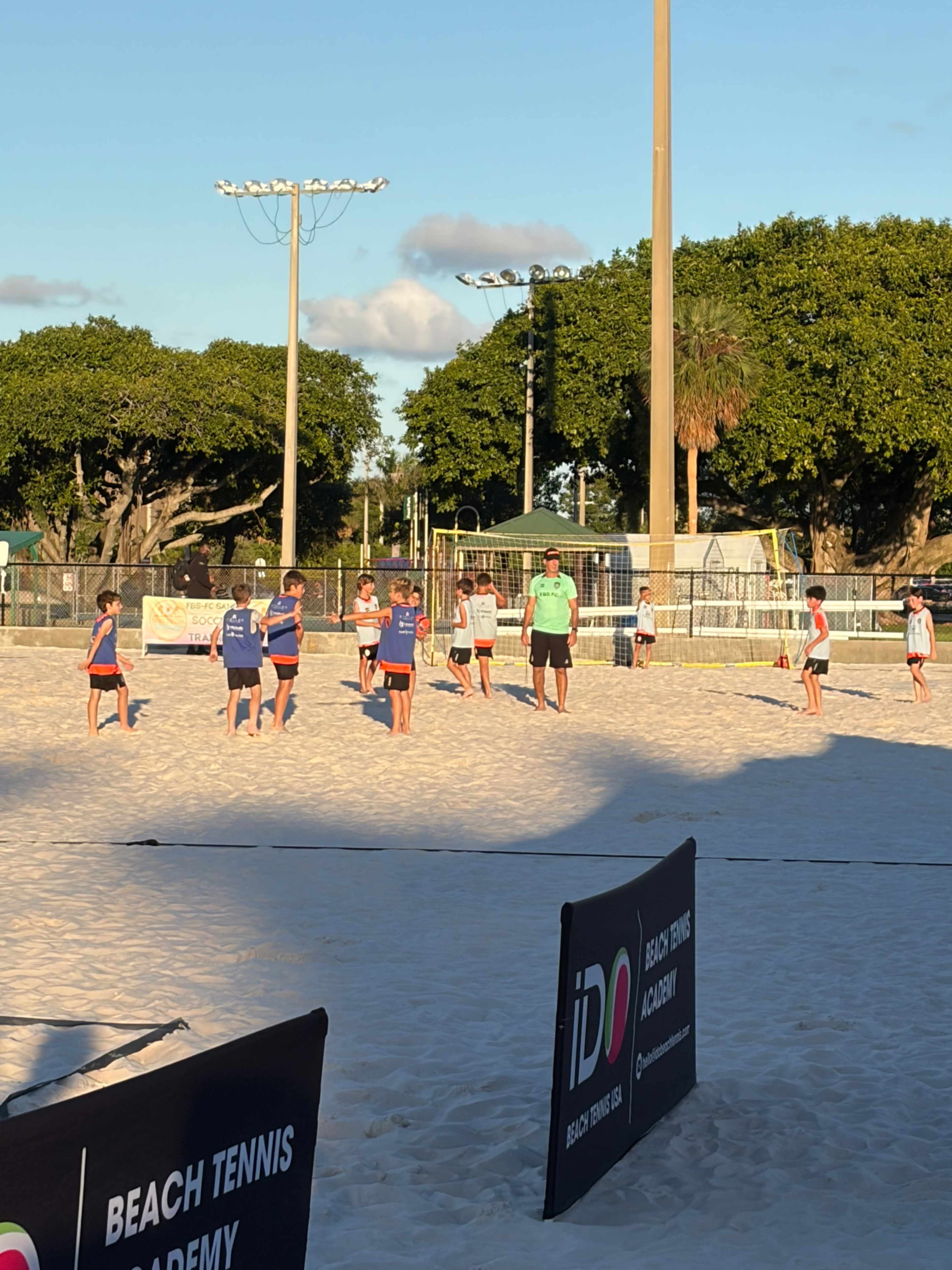 A group of children participates in a beach tennis practice session on a sandy court, with a coach supervising in the background.