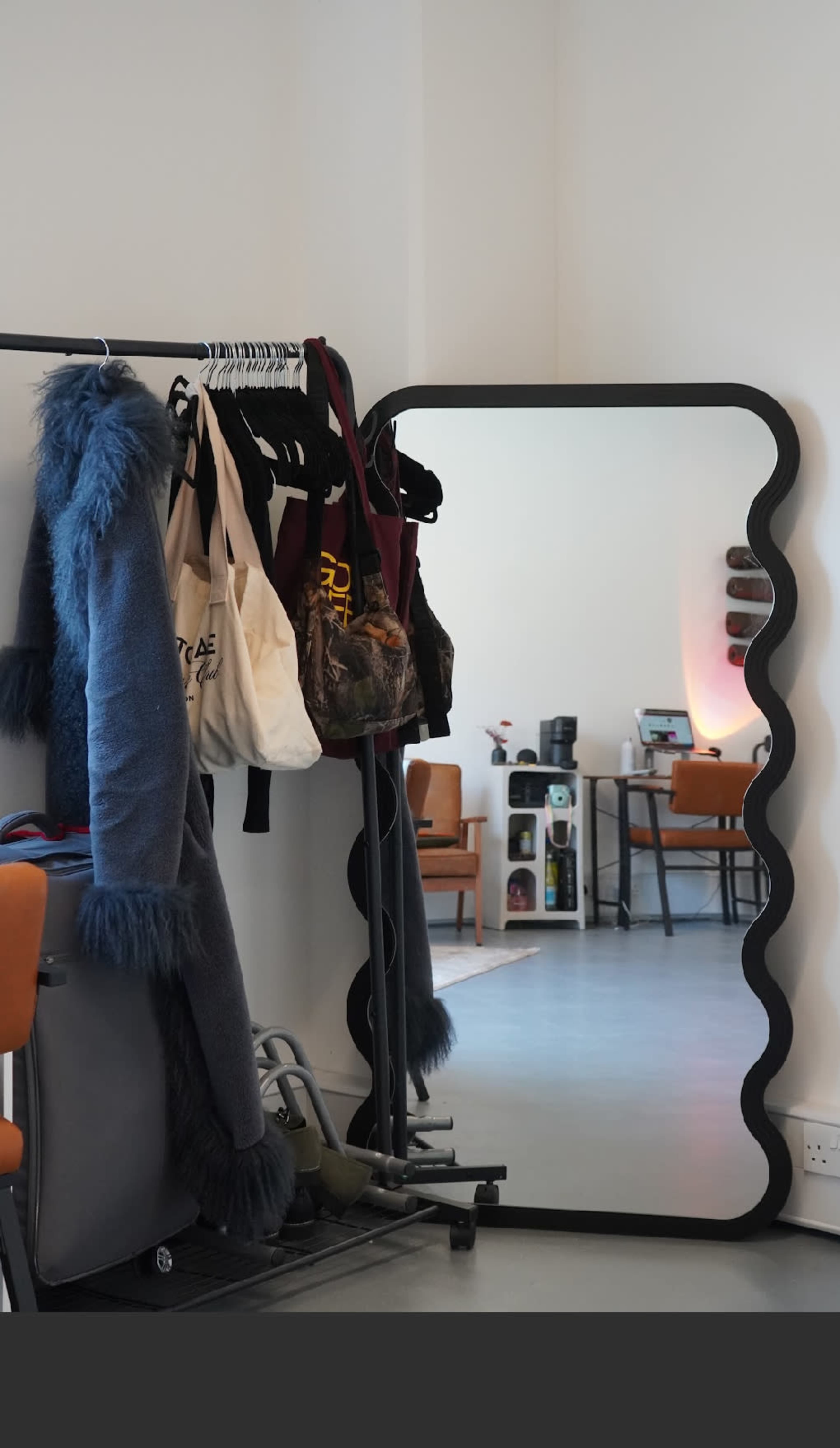 A large, scalloped-edge mirror reflects a room with chairs and a clothing rack holding various garments.