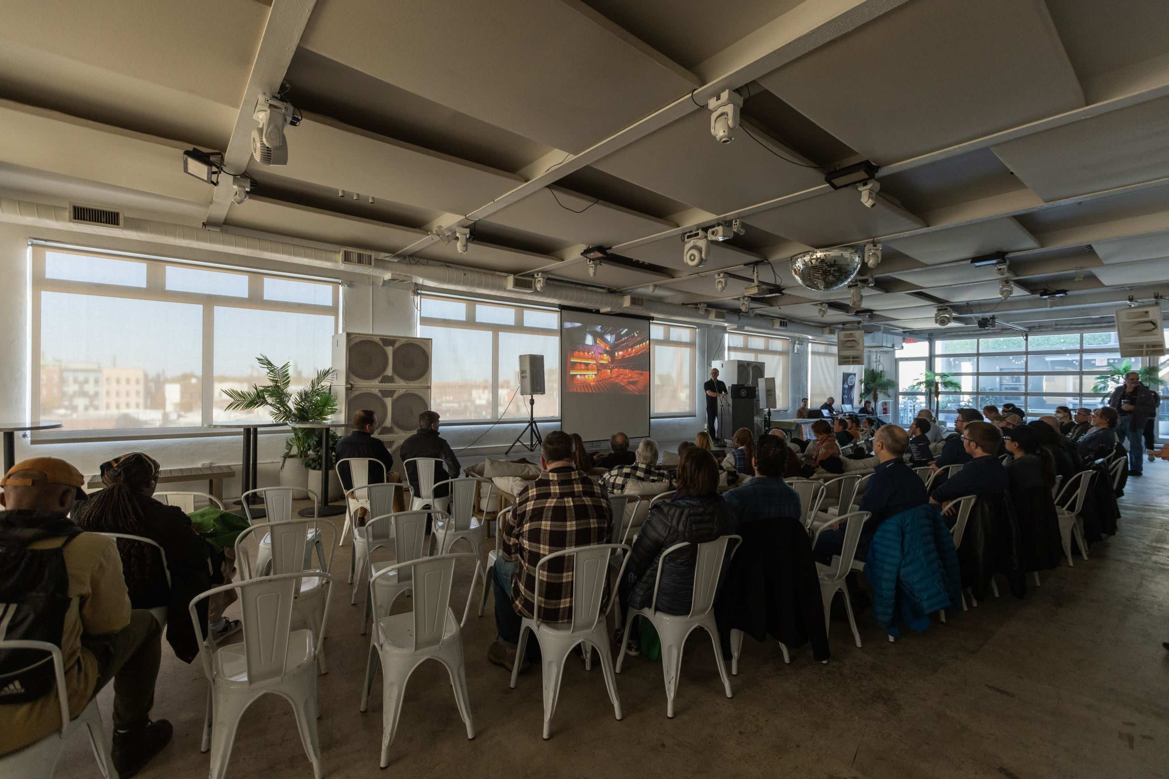 A group of attendees sits in metal chairs, facing a speaker and a large screen displaying visuals in a well-lit room with large windows.