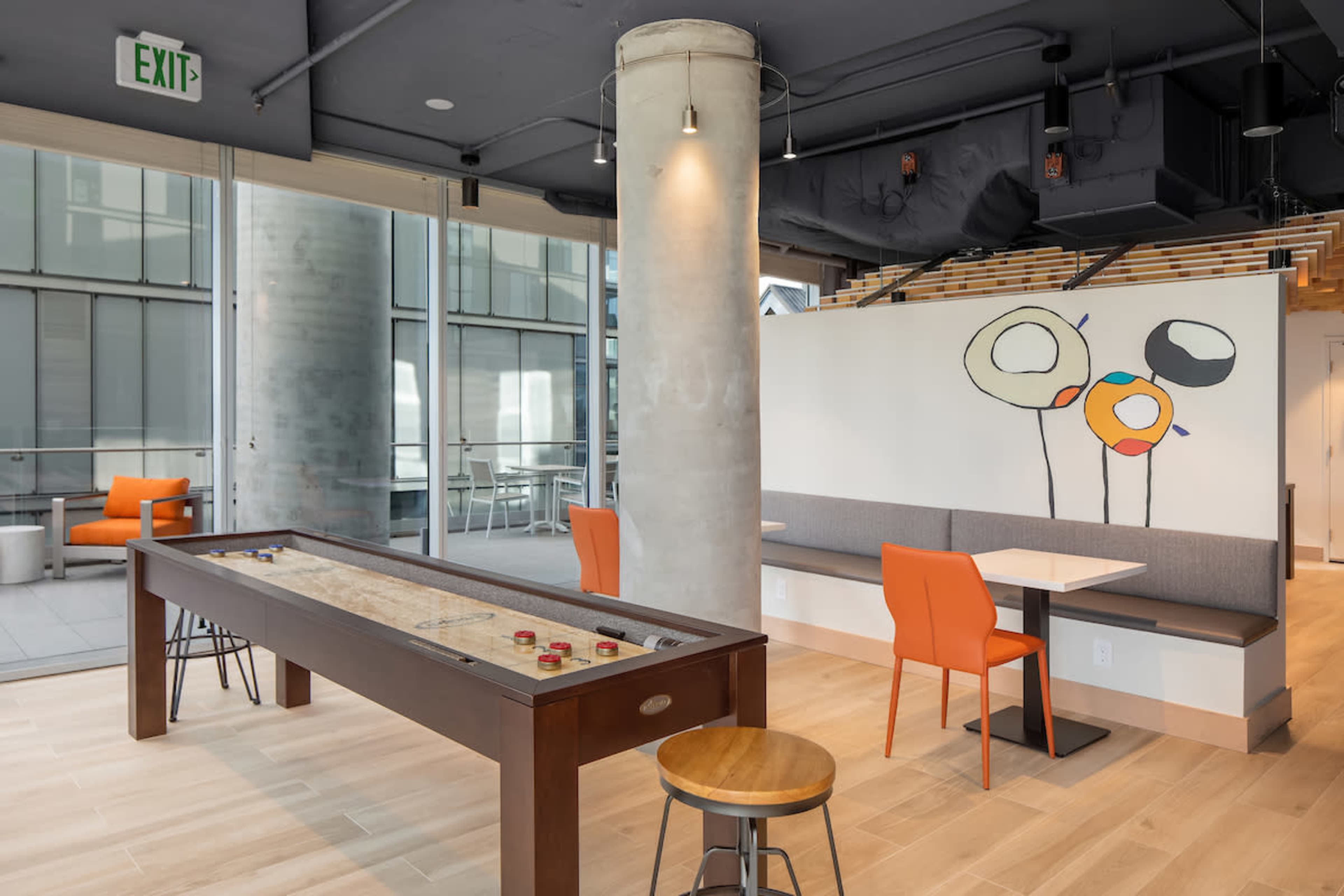 A modern indoor space featuring a long wooden game table, a seating area with a gray bench, orange chairs, and a mural with abstract flower designs.