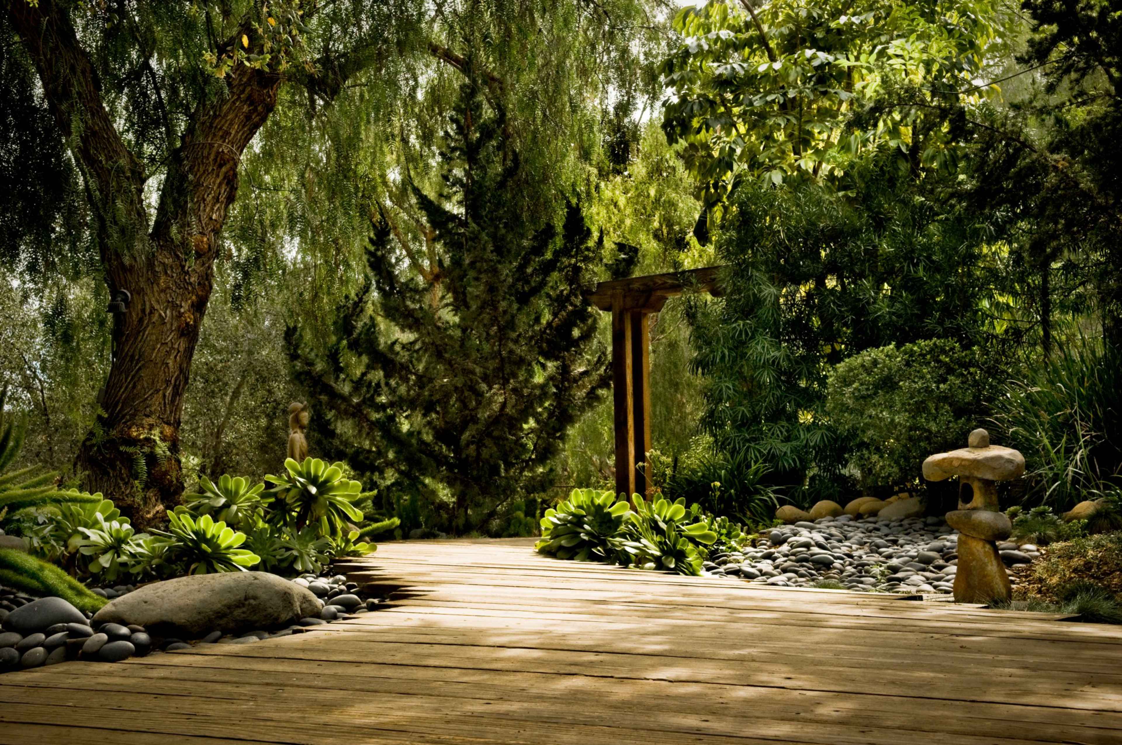 A wooden pathway leads through a landscaped garden featuring stones, lush greenery, and ornamental rocks.