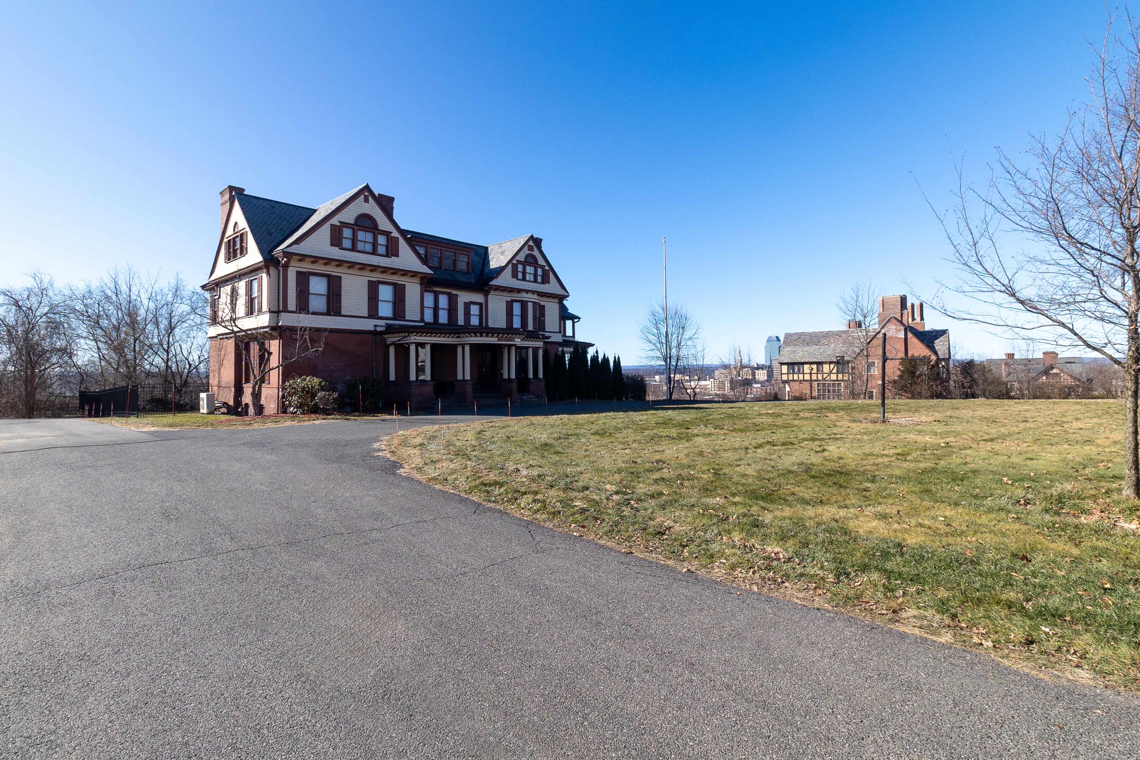 A large, historic brick mansion stands on a winding driveway, surrounded by grassy areas and trees.