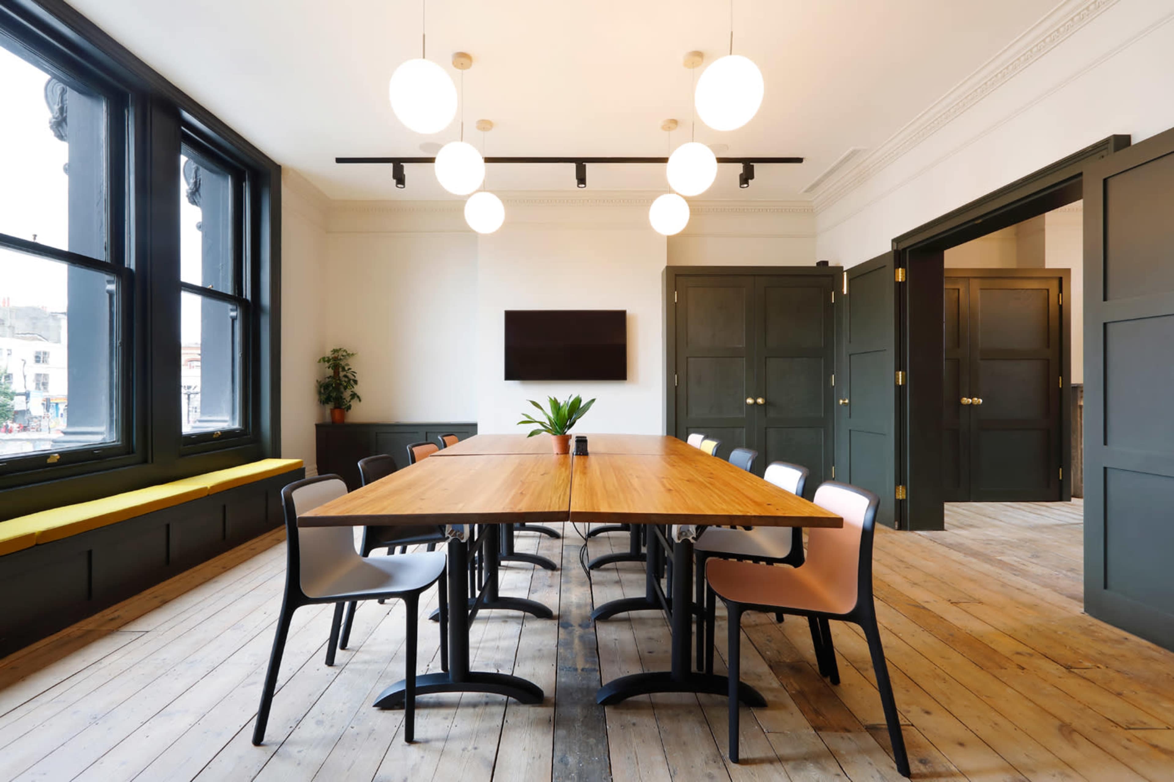 A modern conference room featuring a long wooden table surrounded by chairs, large windows, and a television mounted on the wall.
