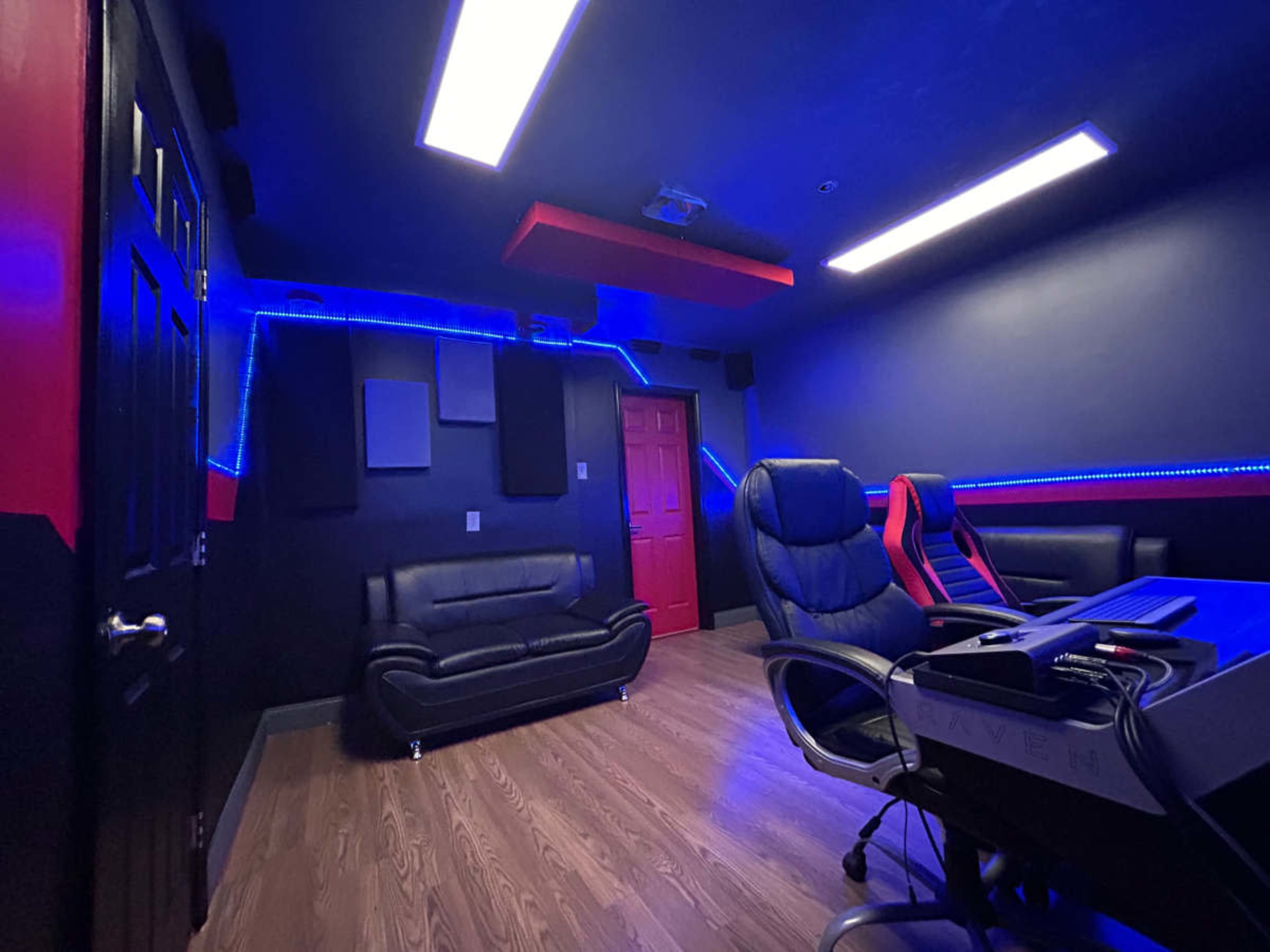 The image shows a modern room featuring black and red walls, a leather sofa, two office chairs, and blue LED lighting.