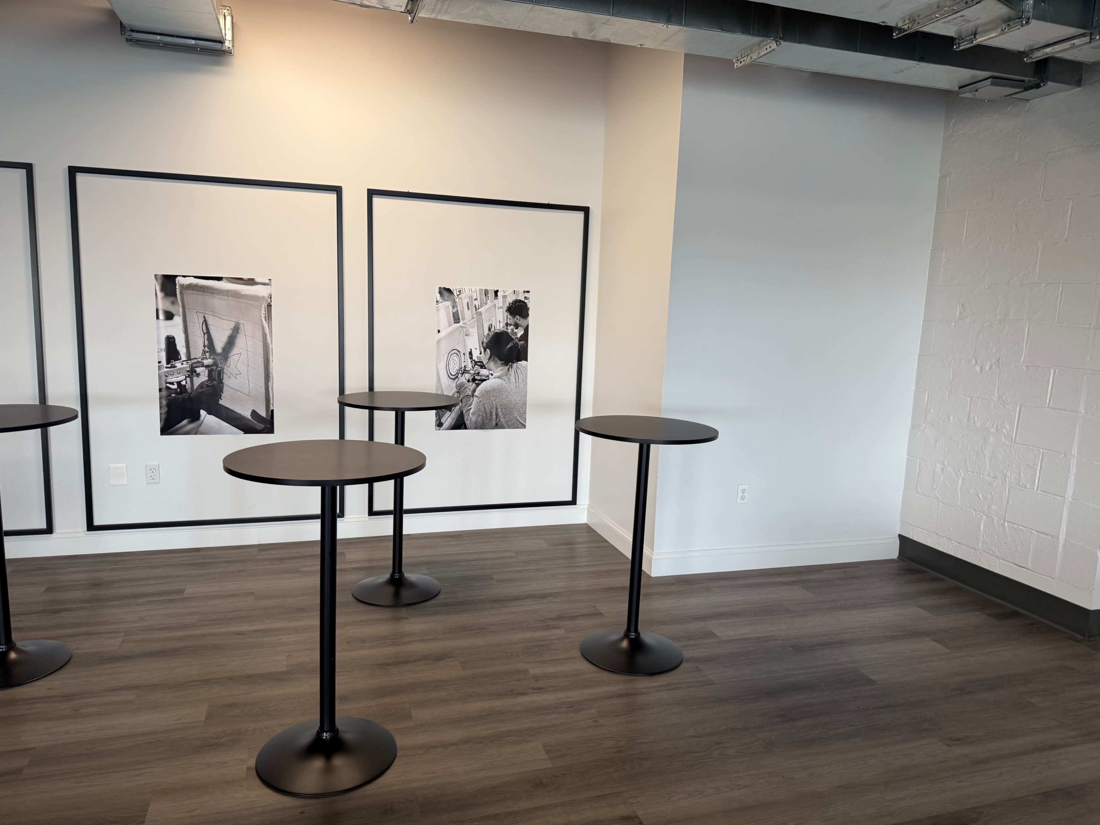 The image shows a room with three tall, round tables arranged near two black-and-white framed photographs on the wall.