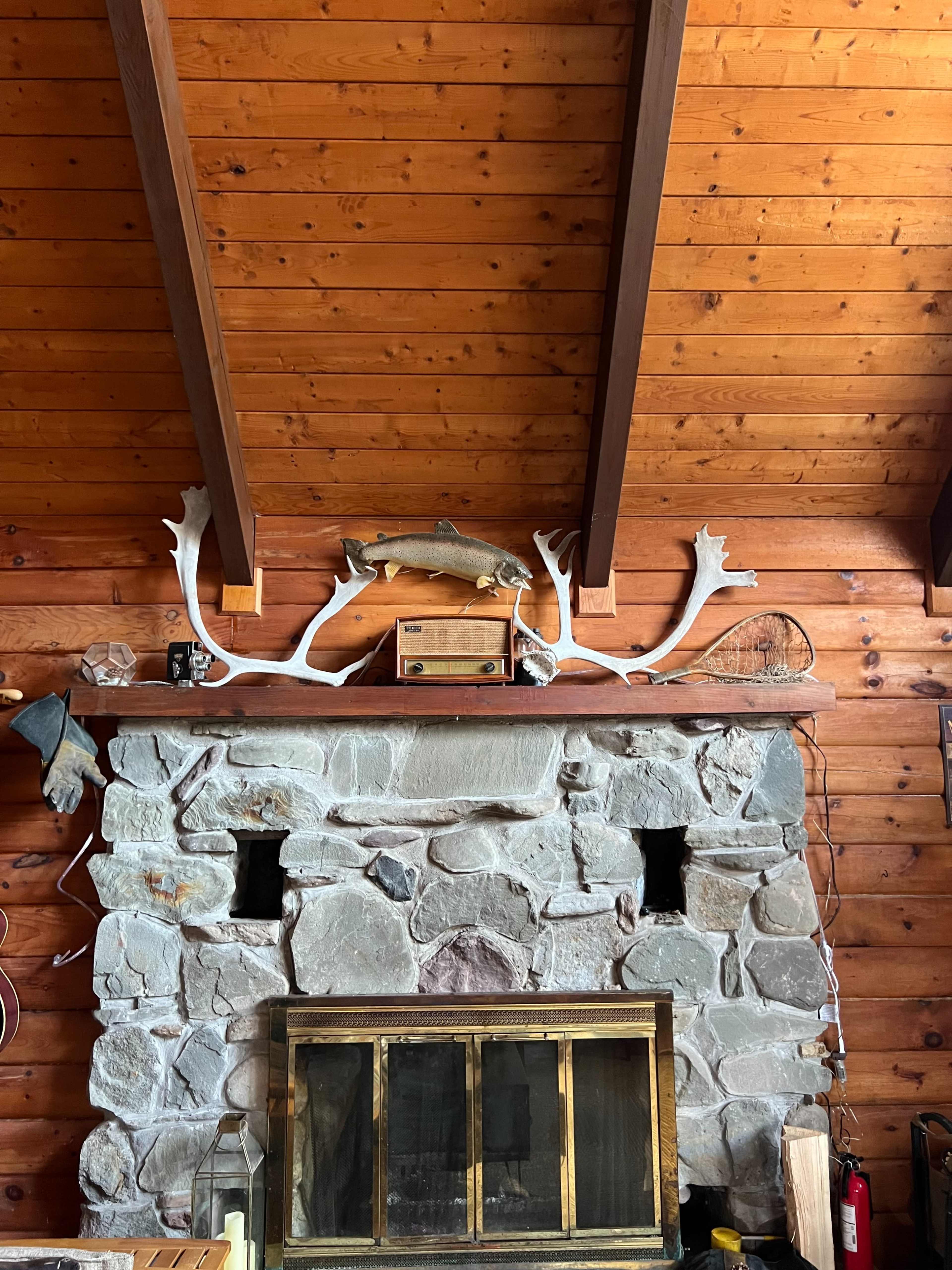 A stone fireplace with a wooden mantel is decorated with a mounted fish, antlers, and various rustic items against a wood-paneled wall.