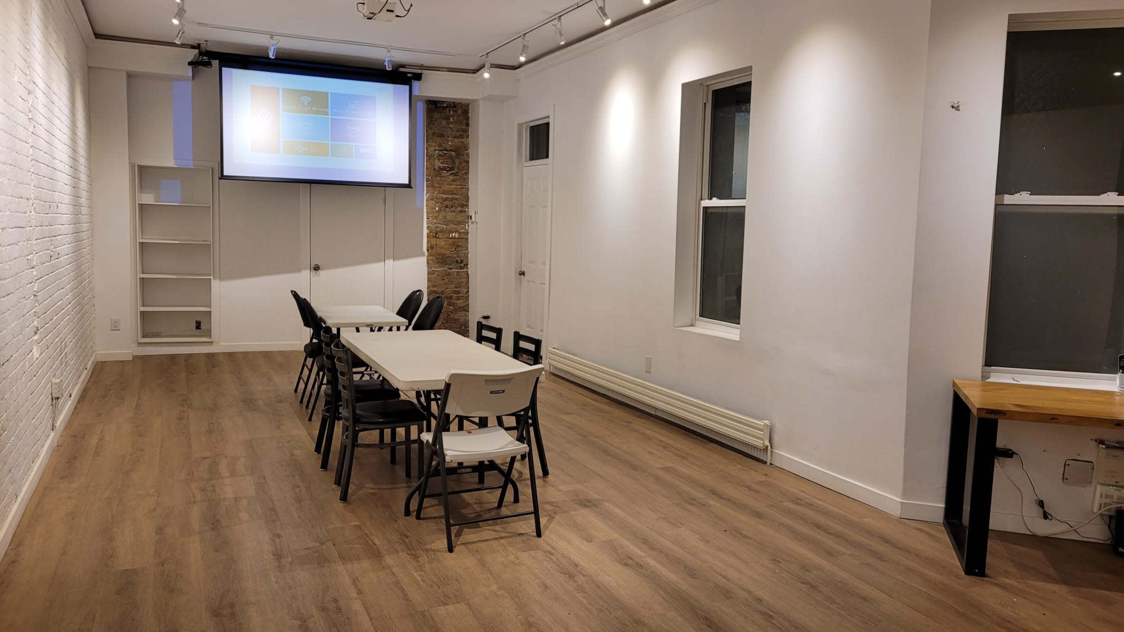 An empty meeting room with a long table surrounded by chairs, a projector screen on the front wall, and windows allowing natural light.
