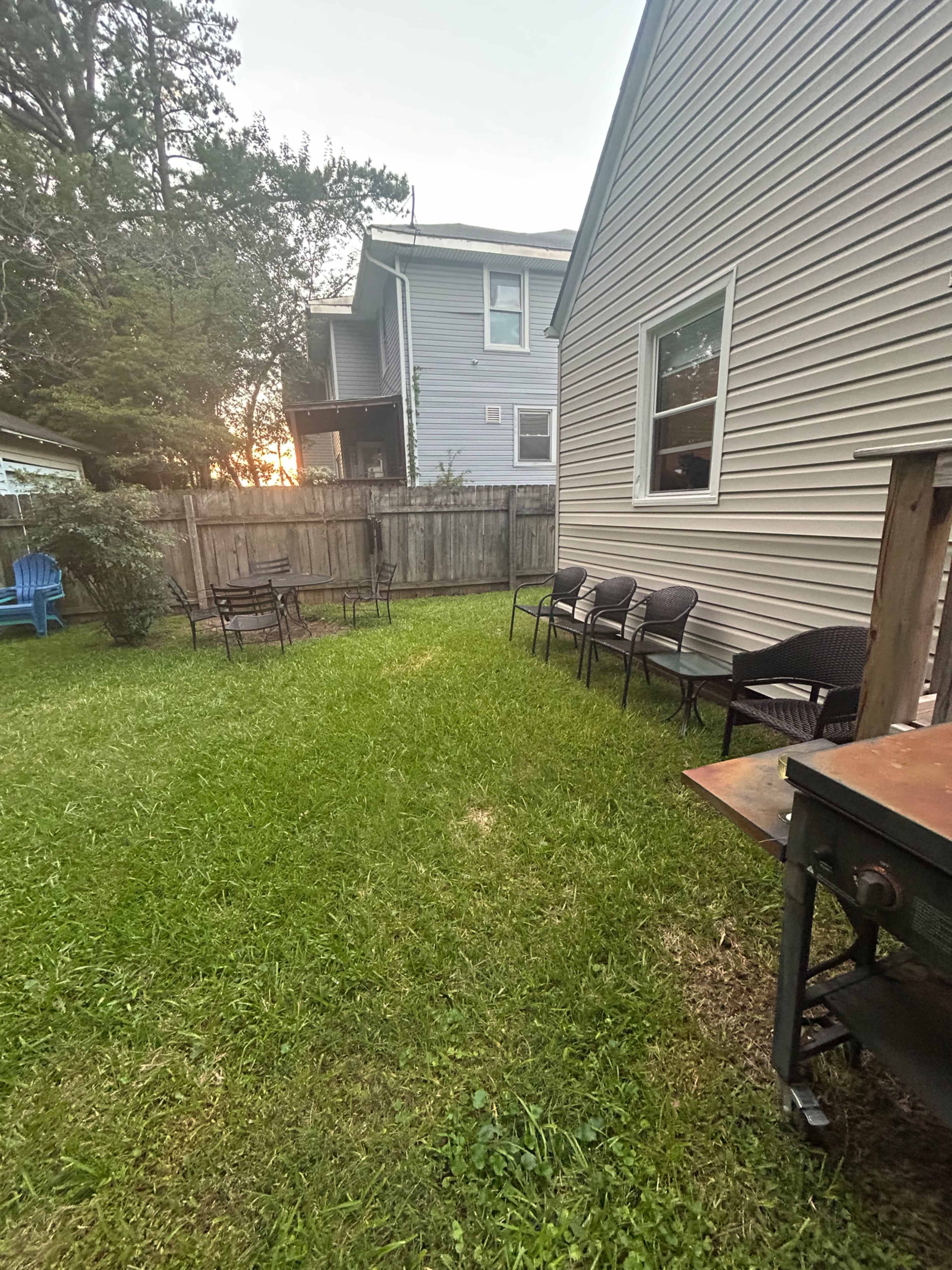 The image shows a grassy backyard area with several black chairs lined up along a fence, a grill on one side, and a glimpse of a sunset through trees in the background.