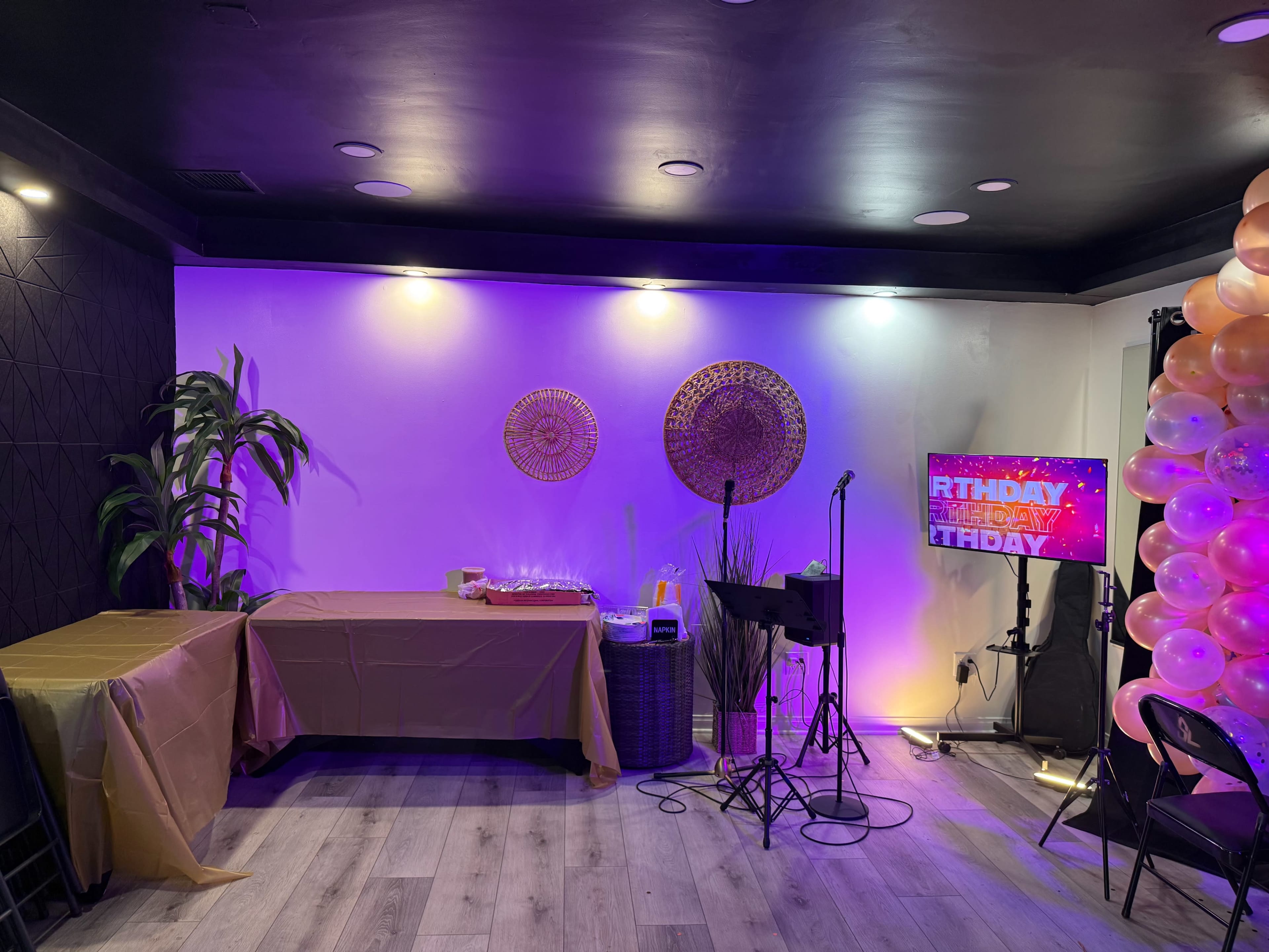 The image shows a party setup with a decorated table, a stage area with a microphone, and colorful balloon decorations.