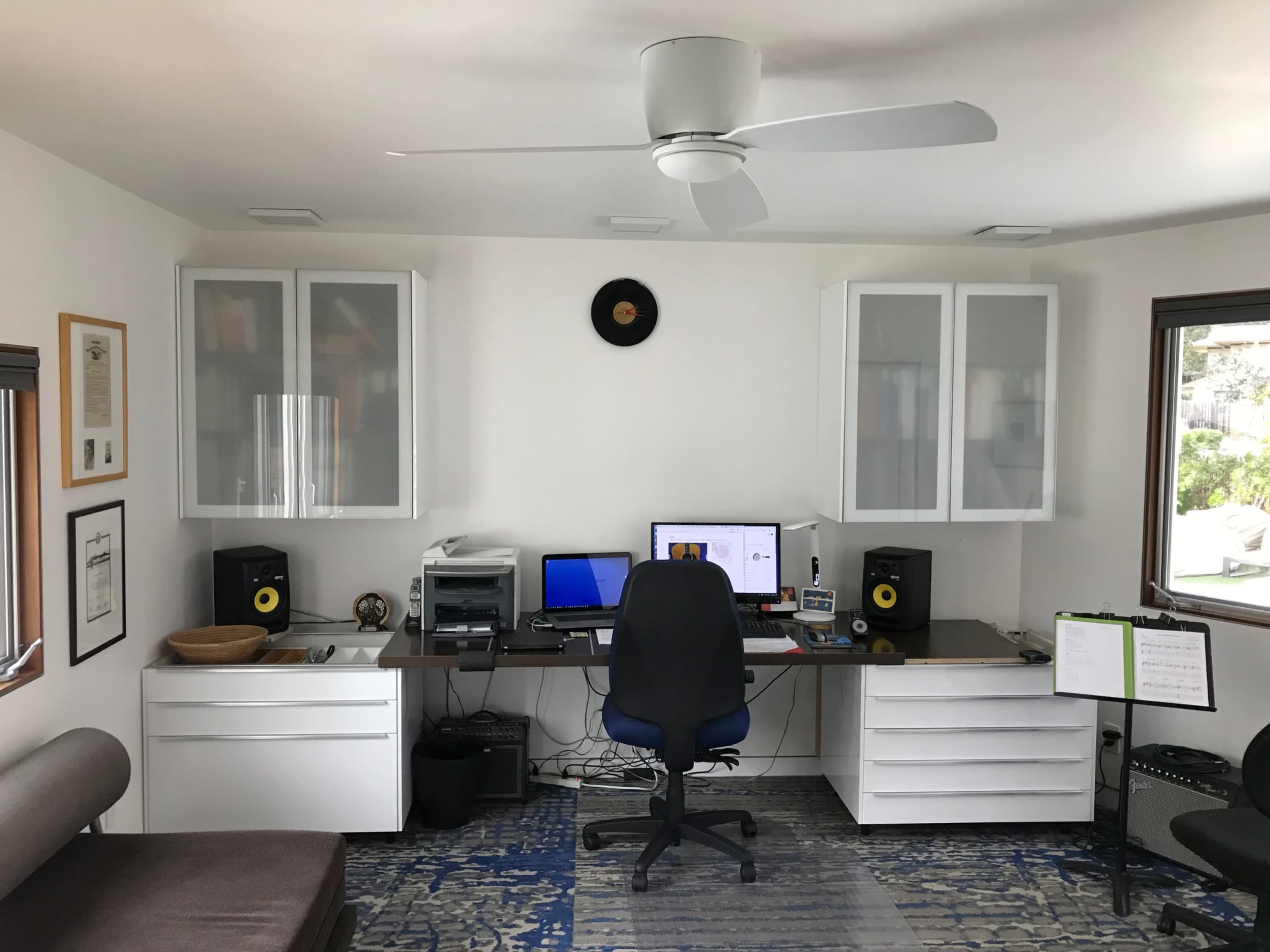 A modern home office with a desk, computer setup, and storage cabinets, flanked by a chair and a music stand.