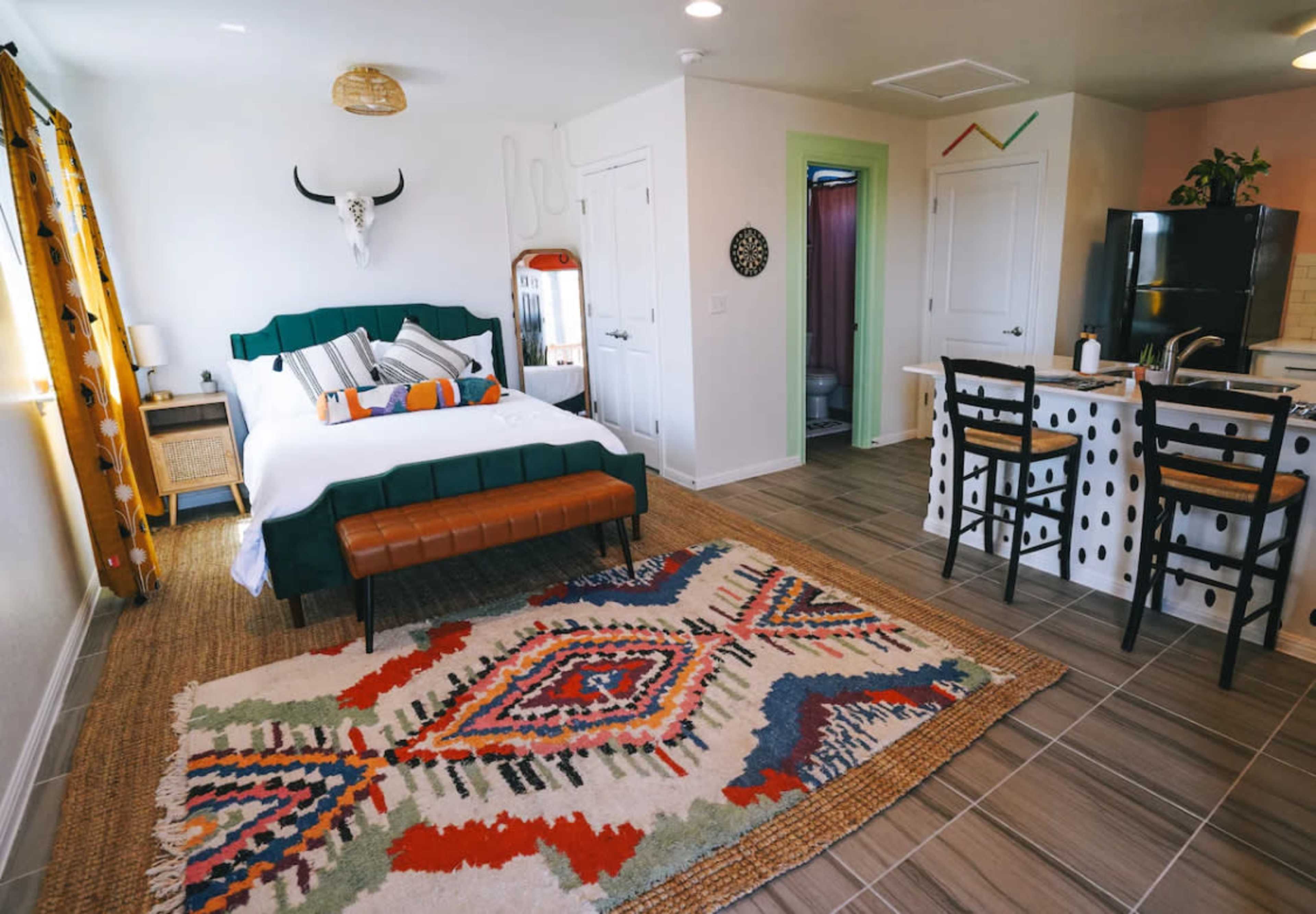 A modern studio apartment features a bed with colorful pillows, a patterned rug, a small dining area with high chairs, and a kitchenette.