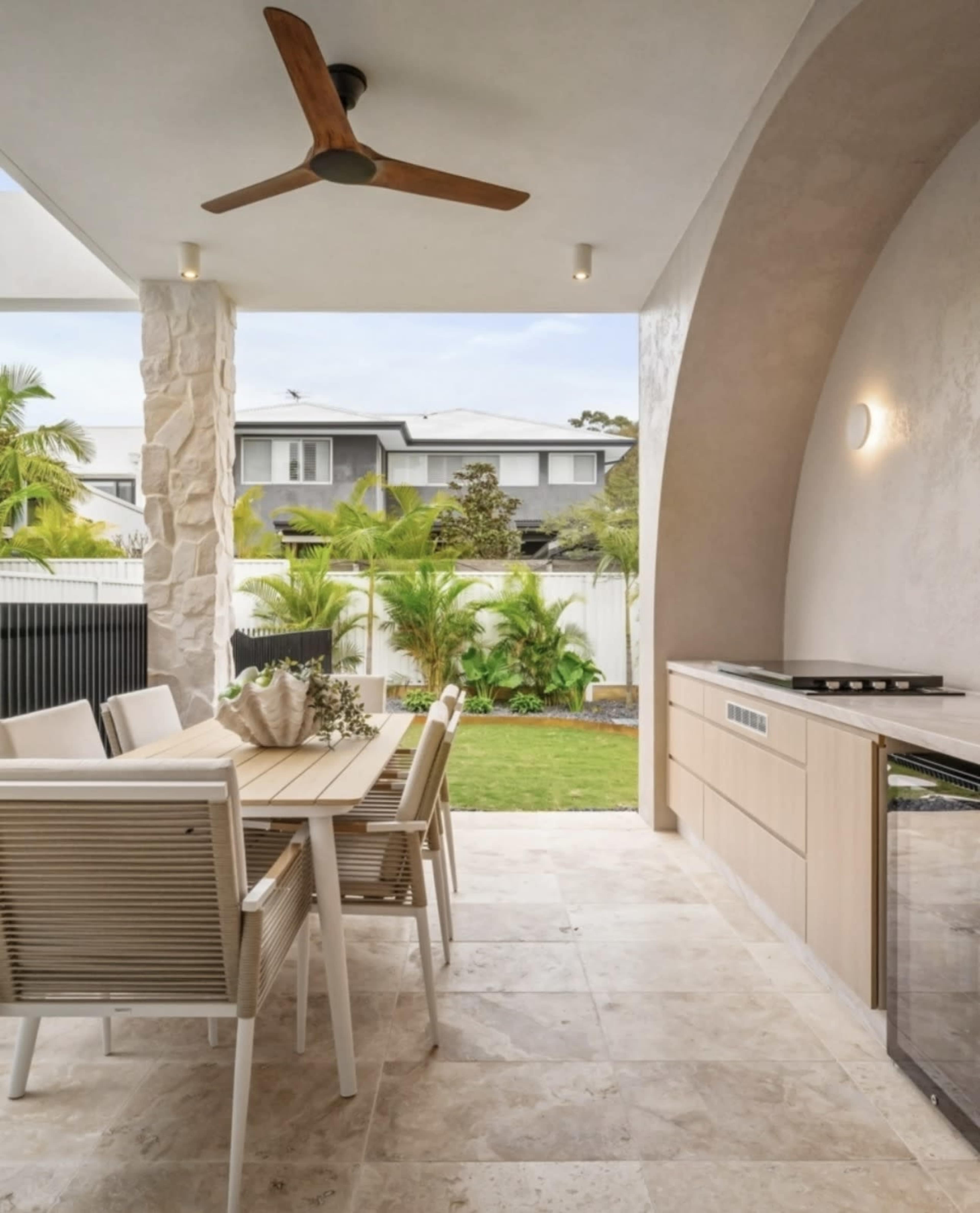 A covered outdoor patio features a dining table and chairs, a grill, and a view of a lush garden surrounded by palm trees and hedges.