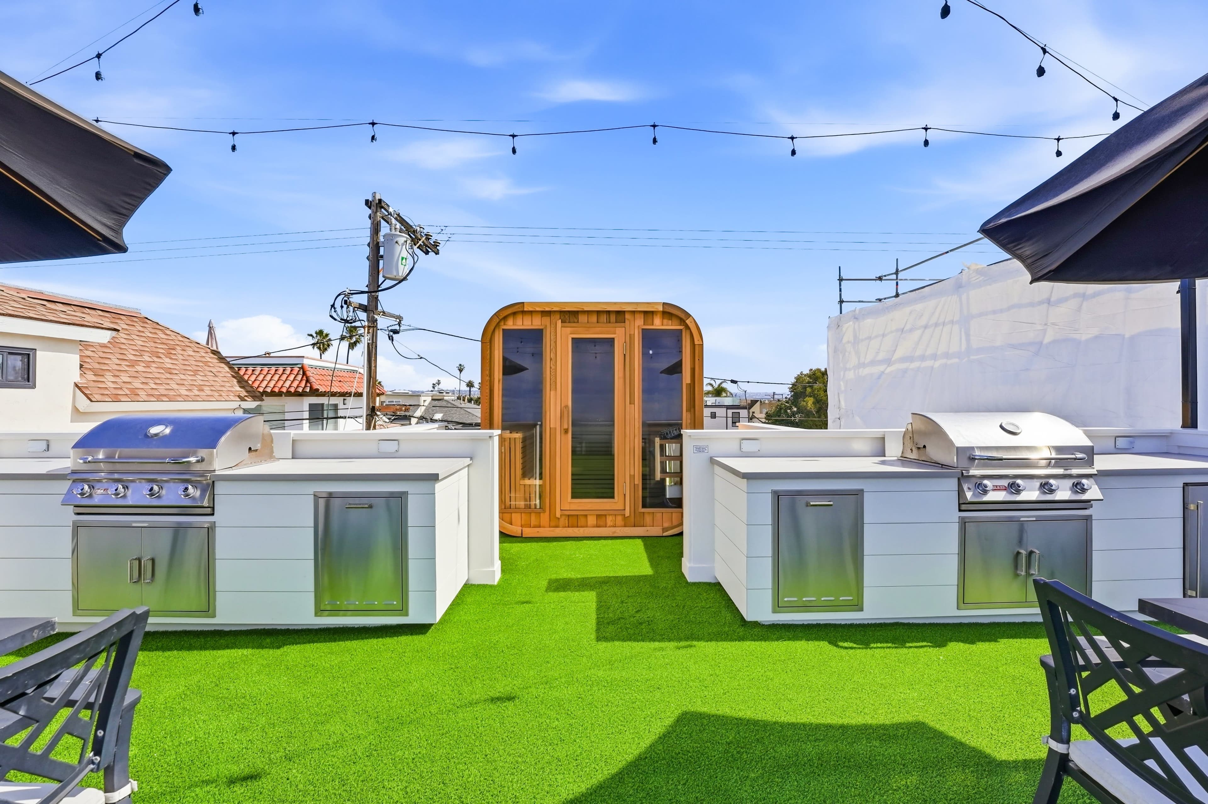 The image shows a rooftop outdoor kitchen with two grill stations and a wooden sauna in the center, all set against a bright blue sky.