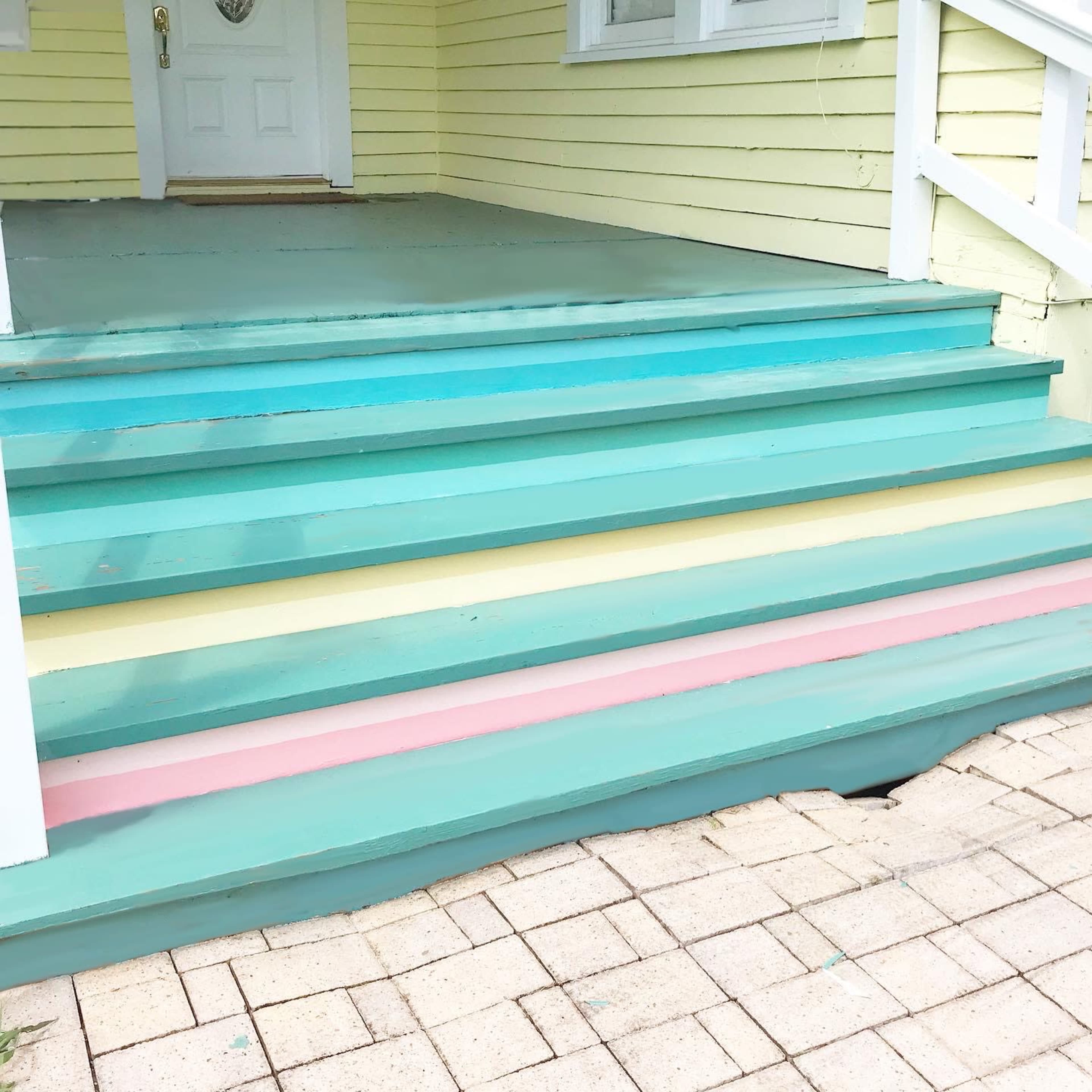 A set of painted wooden steps with alternating turquoise, yellow, and pink stripes, leading up to a door of a house with yellow siding.