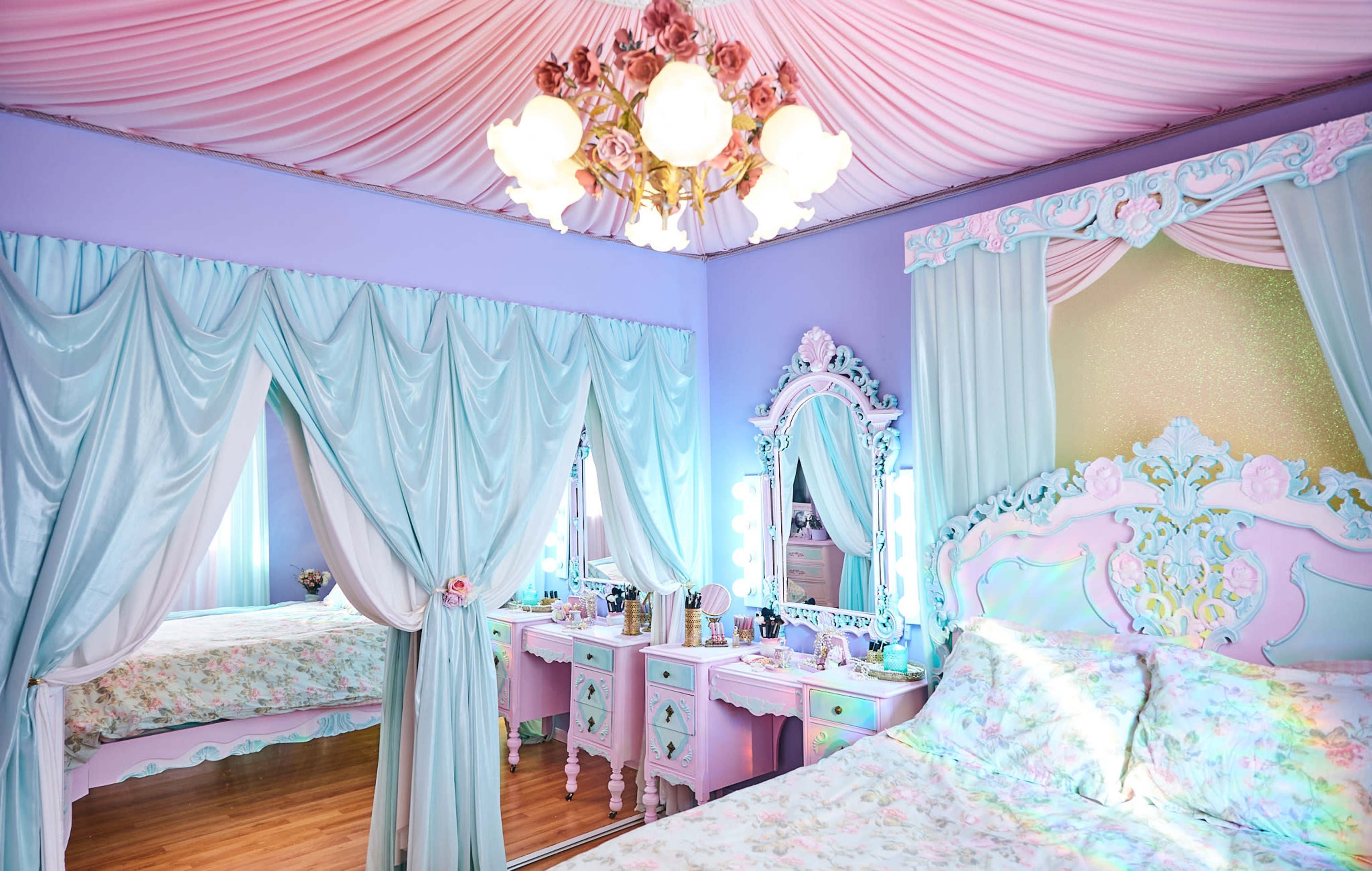 A pastel-colored bedroom with a floral-themed bed, draped curtains, and a vintage-style mirror and vanity set.