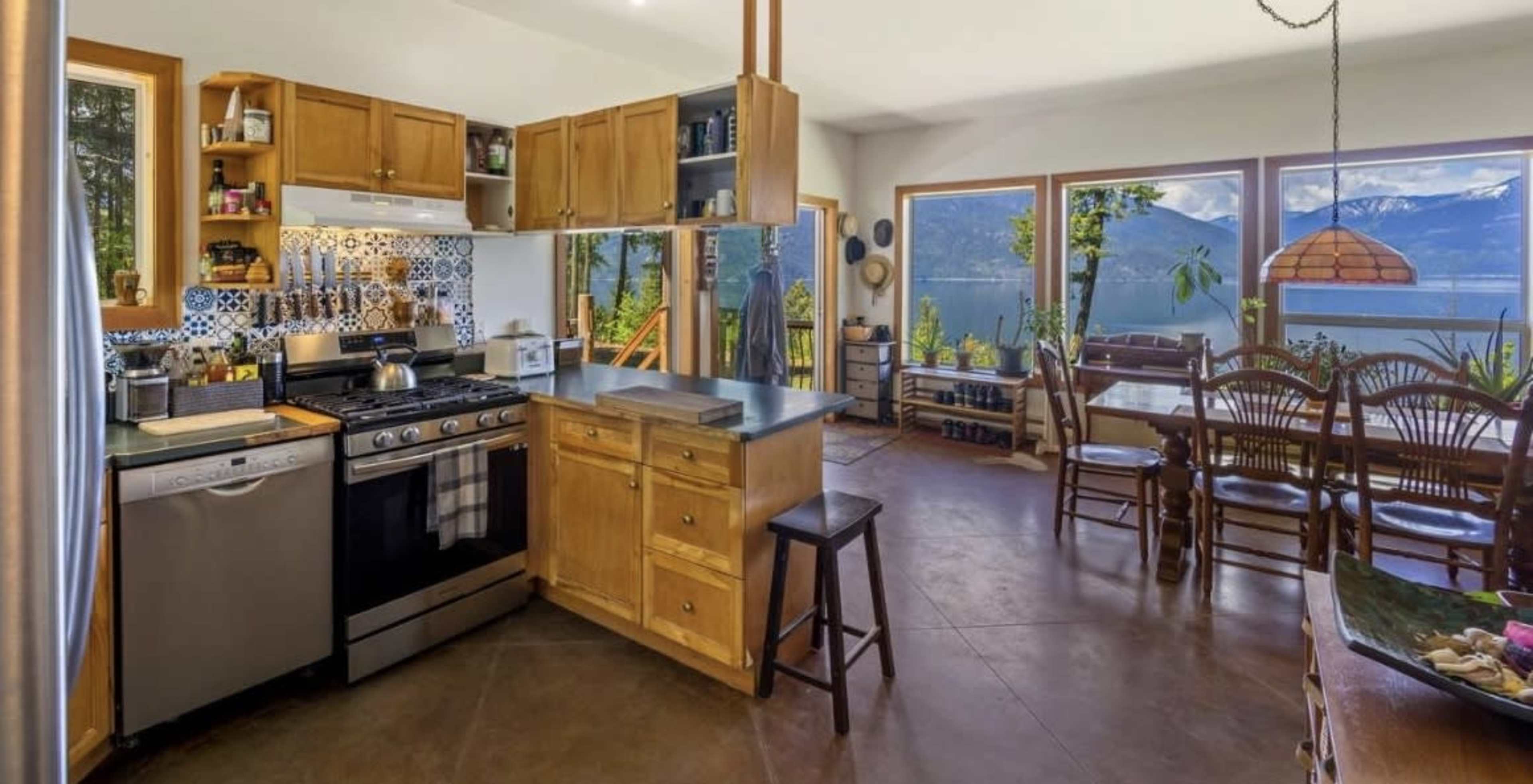 The image shows a modern kitchen with wooden cabinetry, stainless steel appliances, and a dining area featuring large windows with a view of a mountainous landscape.