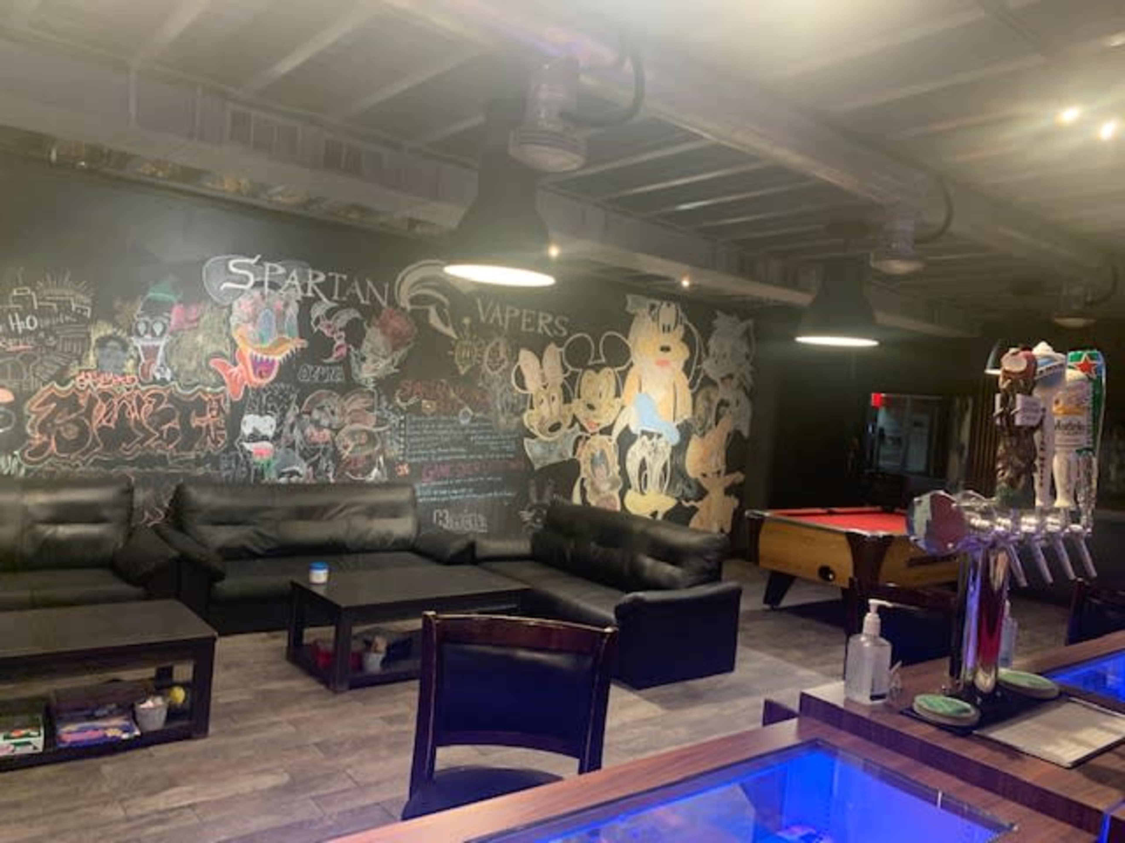 The interior of a lounge features black leather couches, a chalkboard wall filled with graffiti, and a pool table in a dimly lit environment.