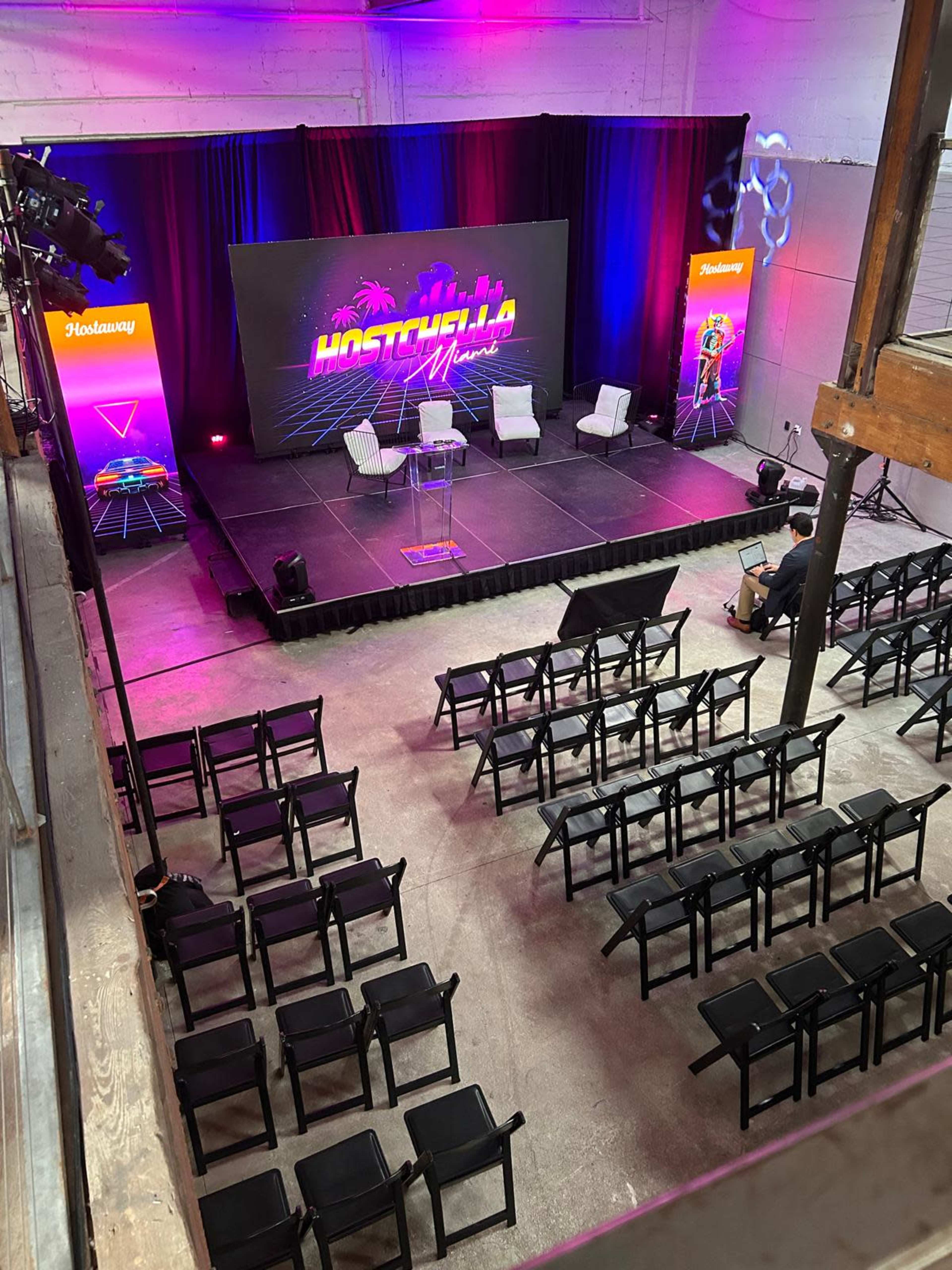 The image shows a stage setup for an event, with a brightly lit backdrop featuring the text "Hosthella," surrounded by empty rows of chairs.