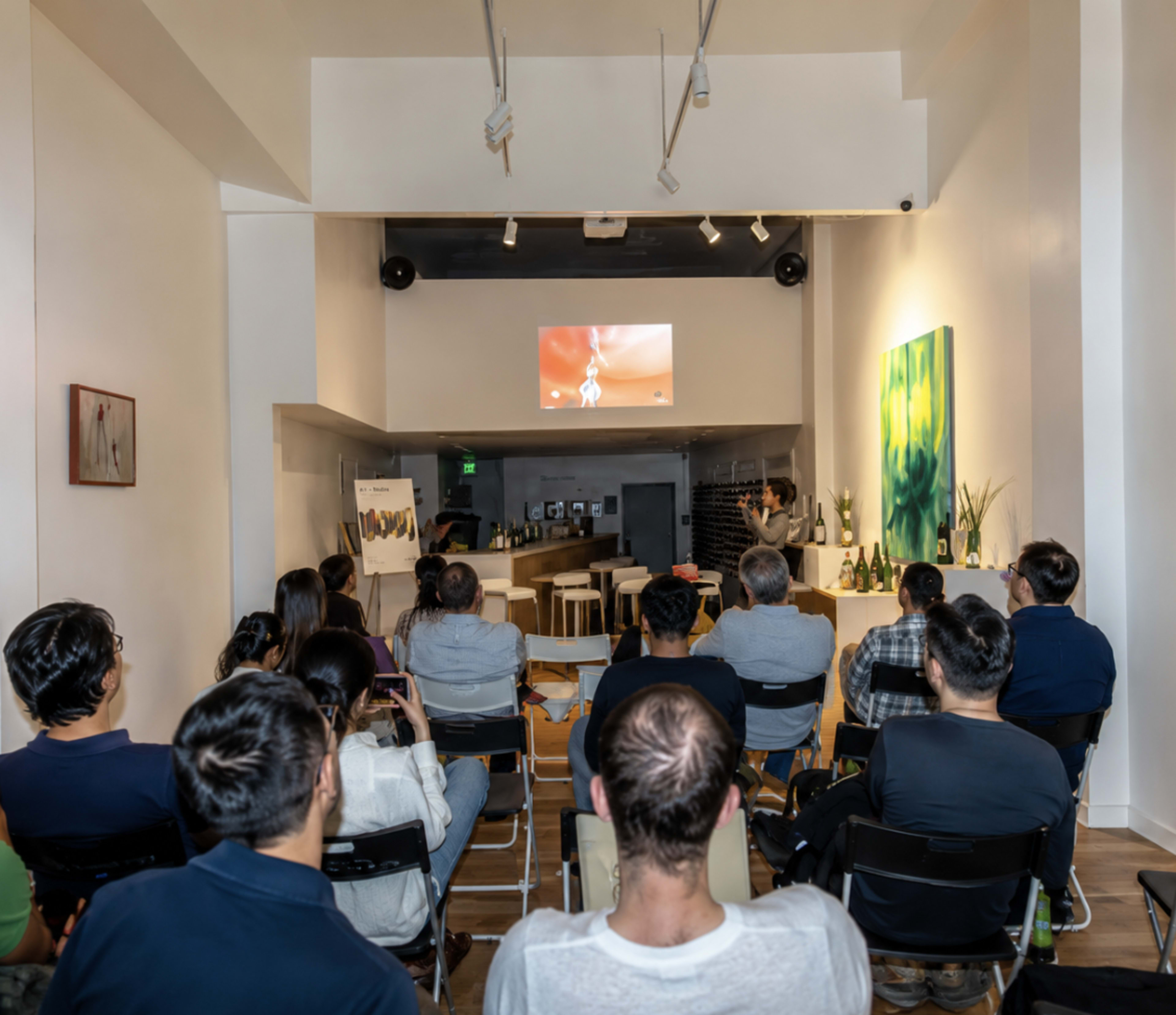 A group of people sits in chairs facing a screen in a modern, bright room with a bar and art on the walls.