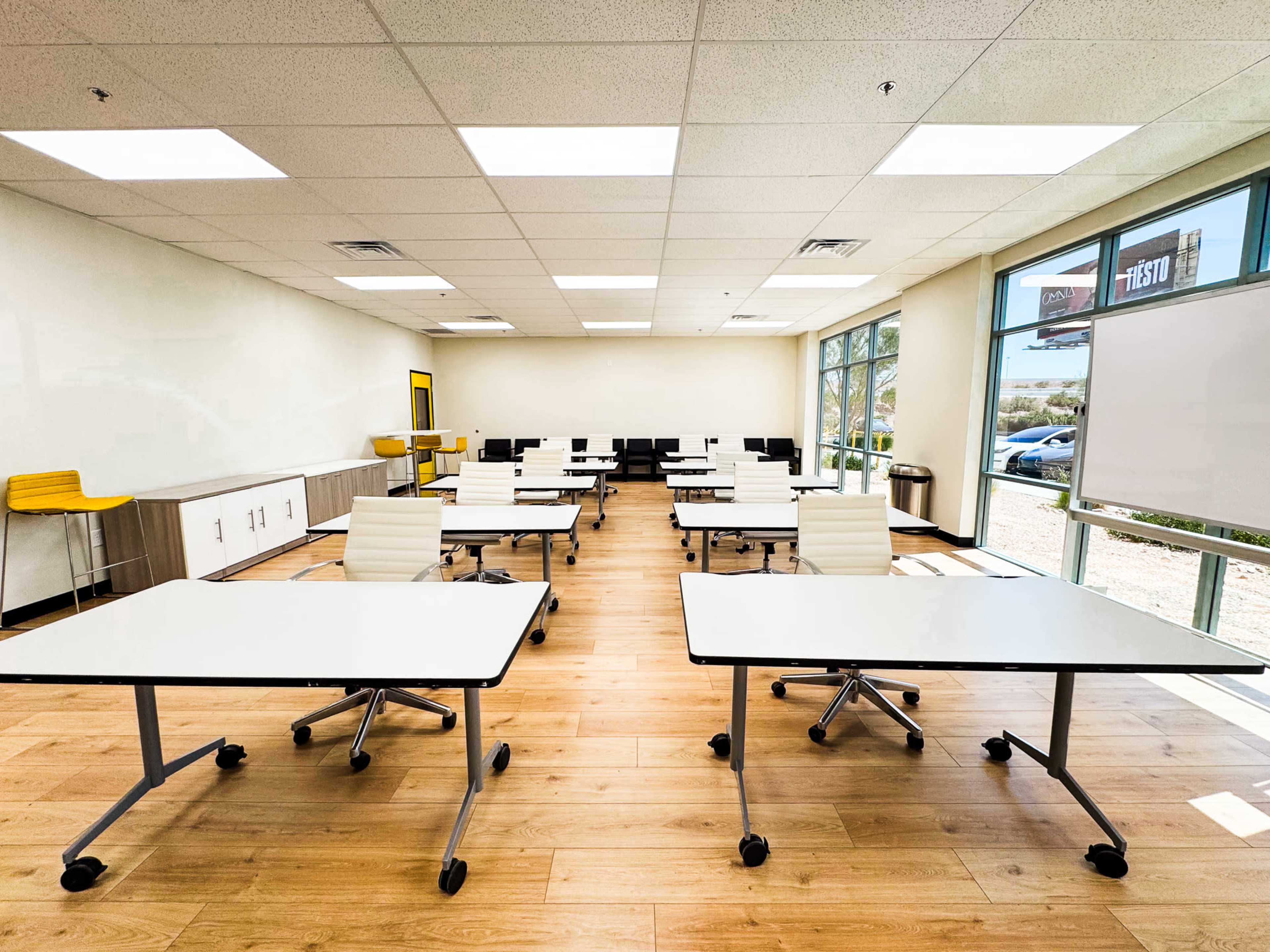 The image shows a modern classroom with several movable desks arranged in a spacious layout, large windows allowing natural light, and a whiteboard on one wall.