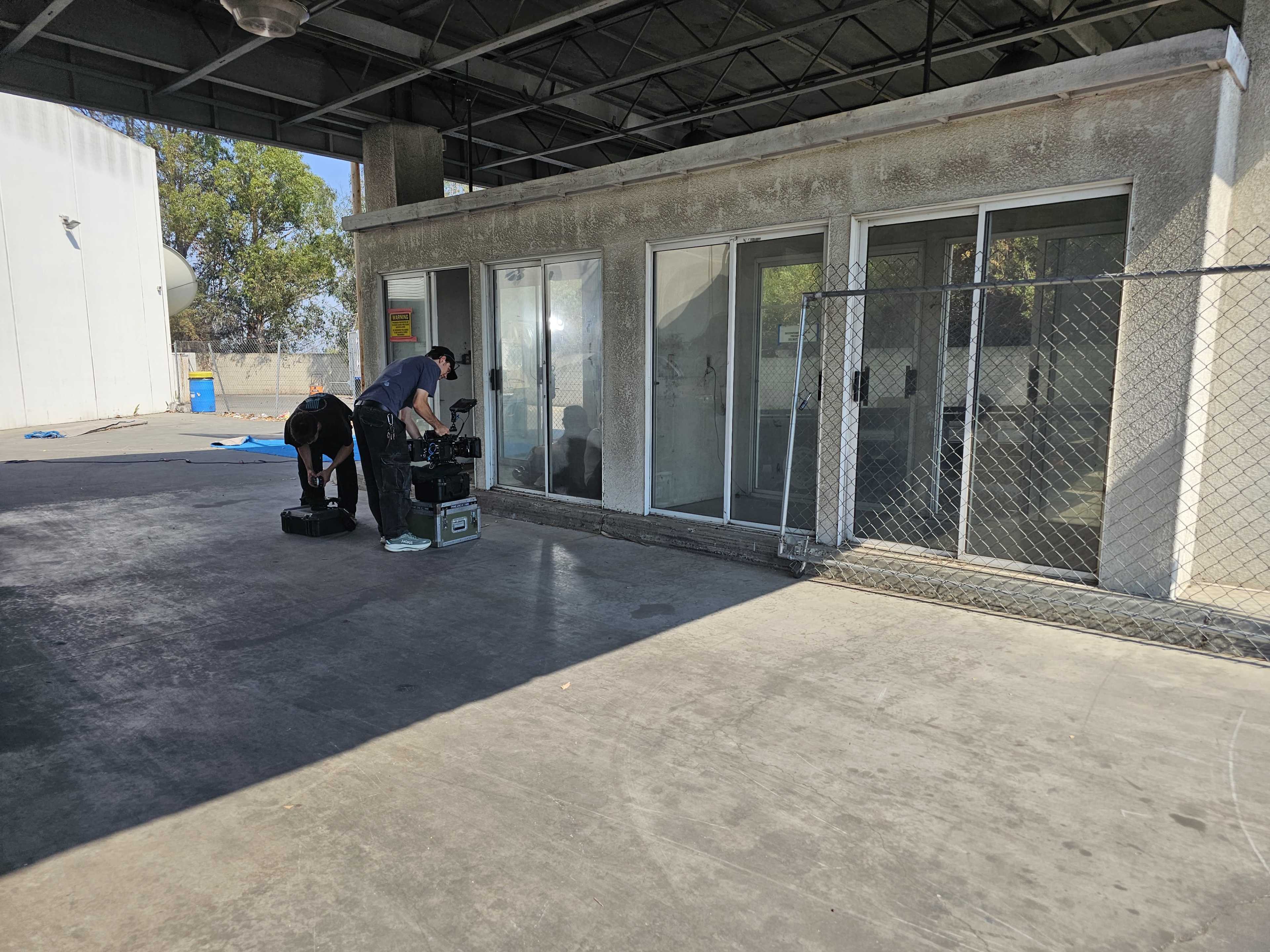 Two individuals are setting up camera equipment in an industrial area next to a building with glass doors and a fence.
