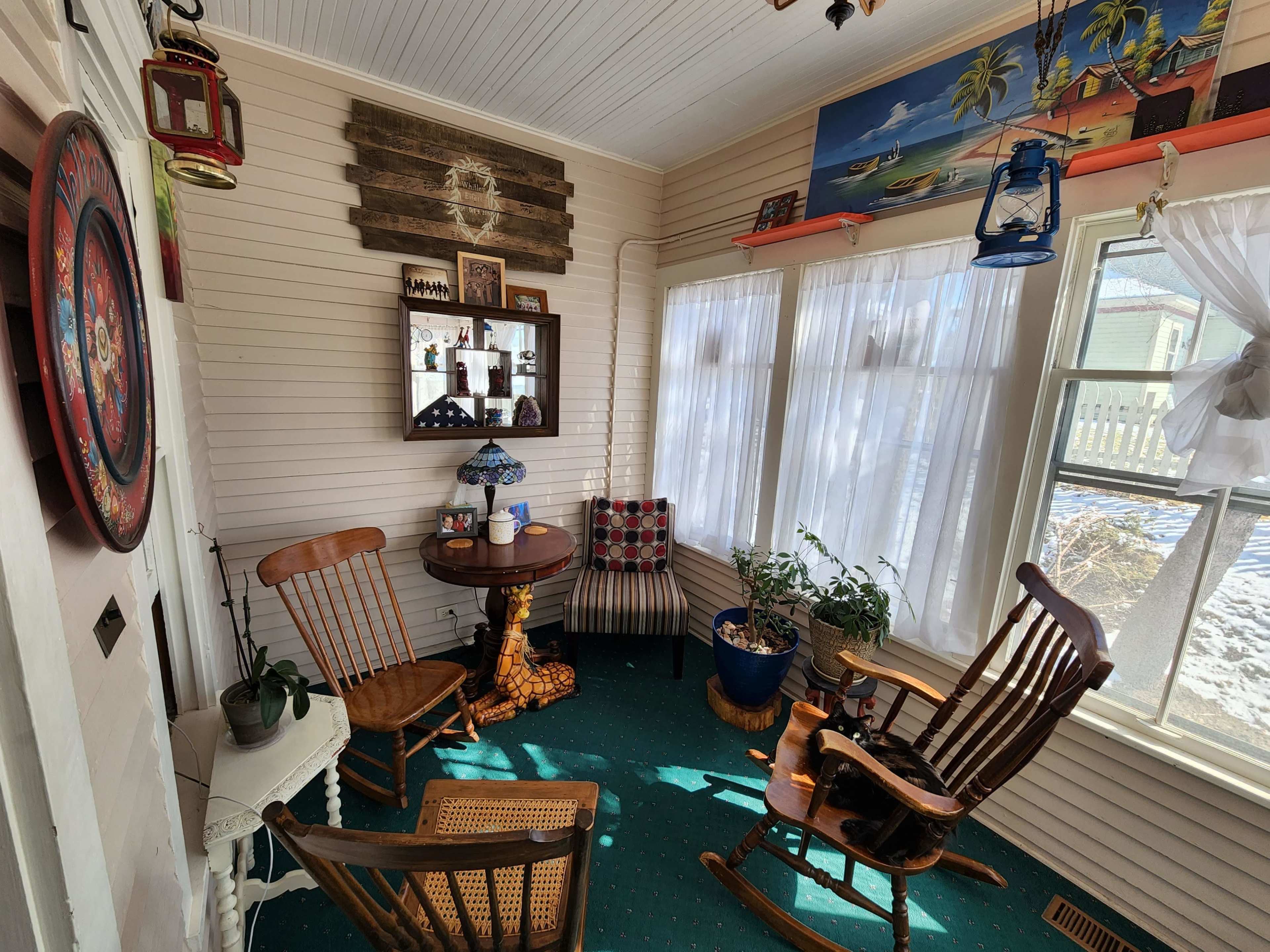 A sunlit sunroom features a small round table surrounded by rocking chairs, with potted plants and decorative items on the walls.