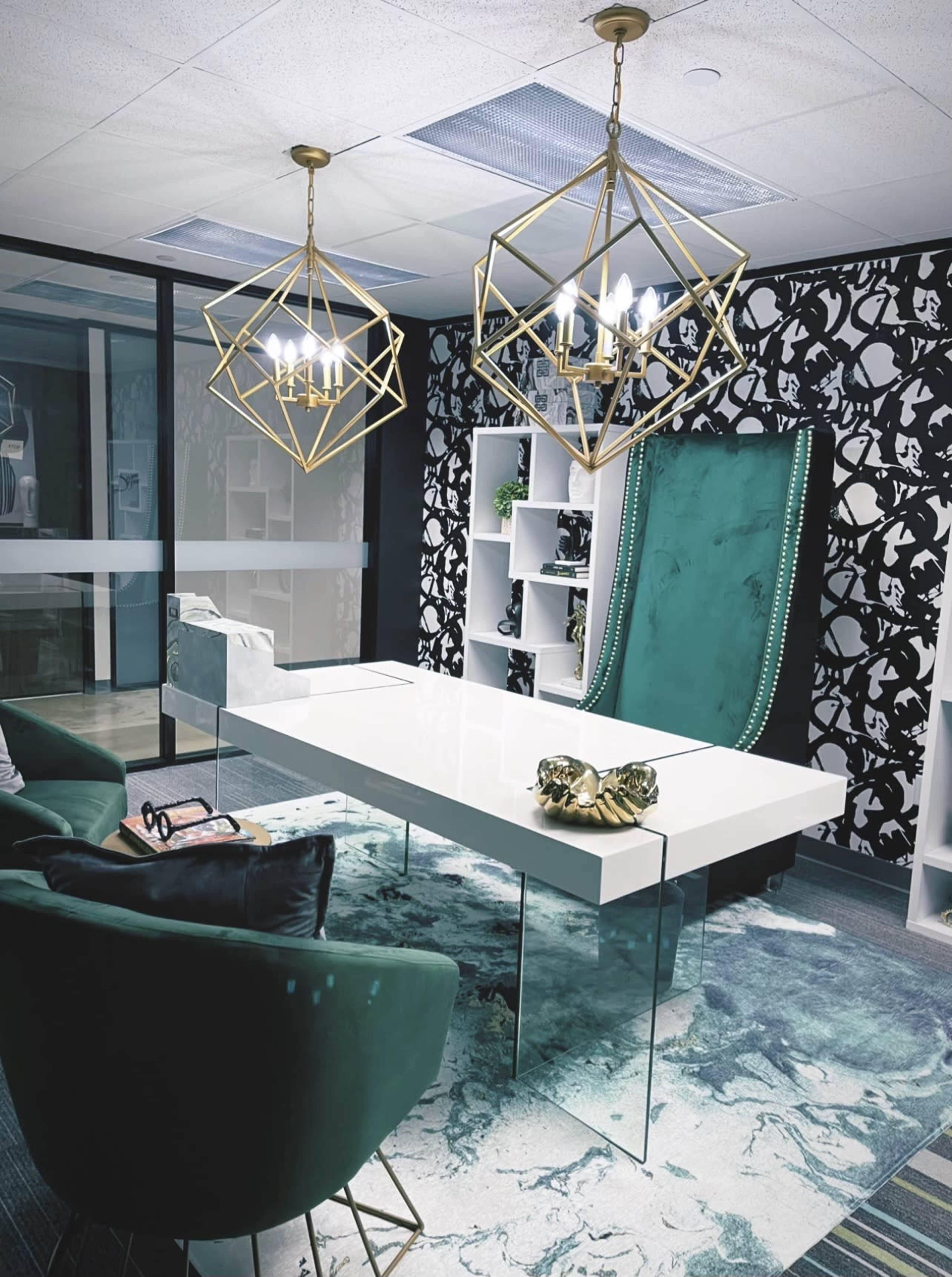 A modern office with a white desk, green velvet-accented chairs, and two geometric gold chandeliers hanging from the ceiling.