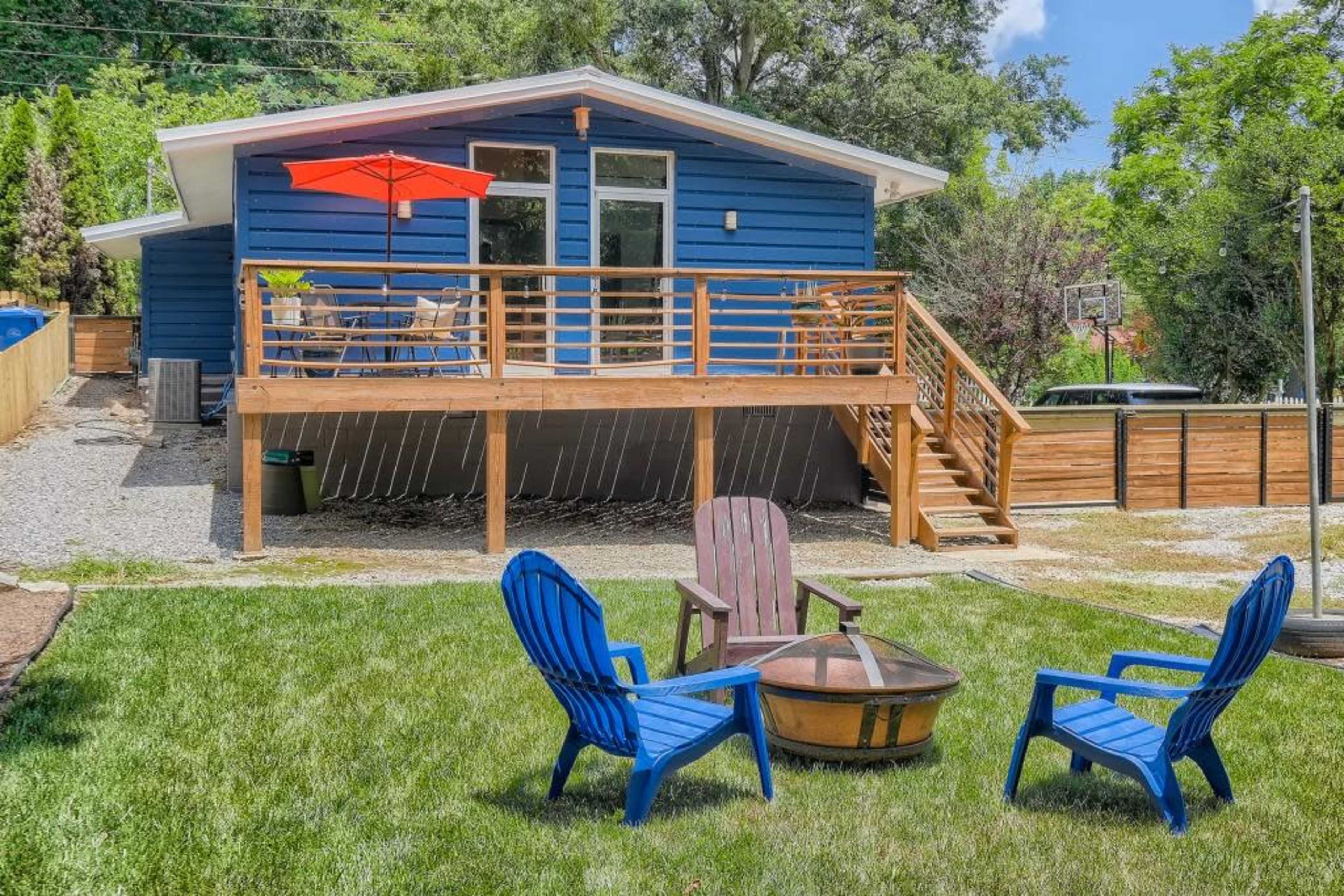A blue house with a wooden deck and red umbrella overlooks a grassy yard, featuring four outdoor chairs arranged around a fire pit.