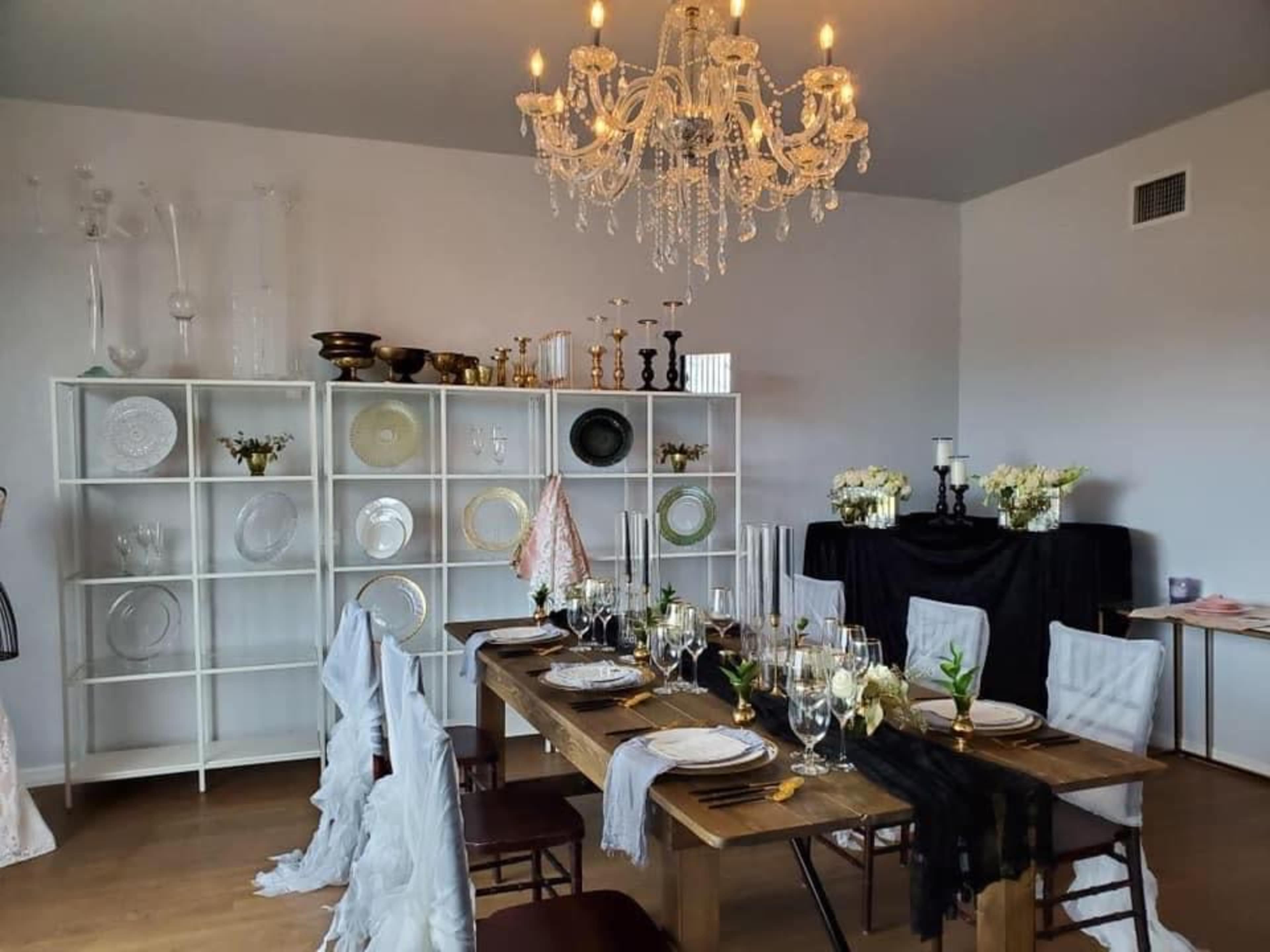 A dining room is set with an elegantly arranged table featuring white plates, glasses, and floral decorations, alongside a display of decorative dishware and a chandelier overhead.