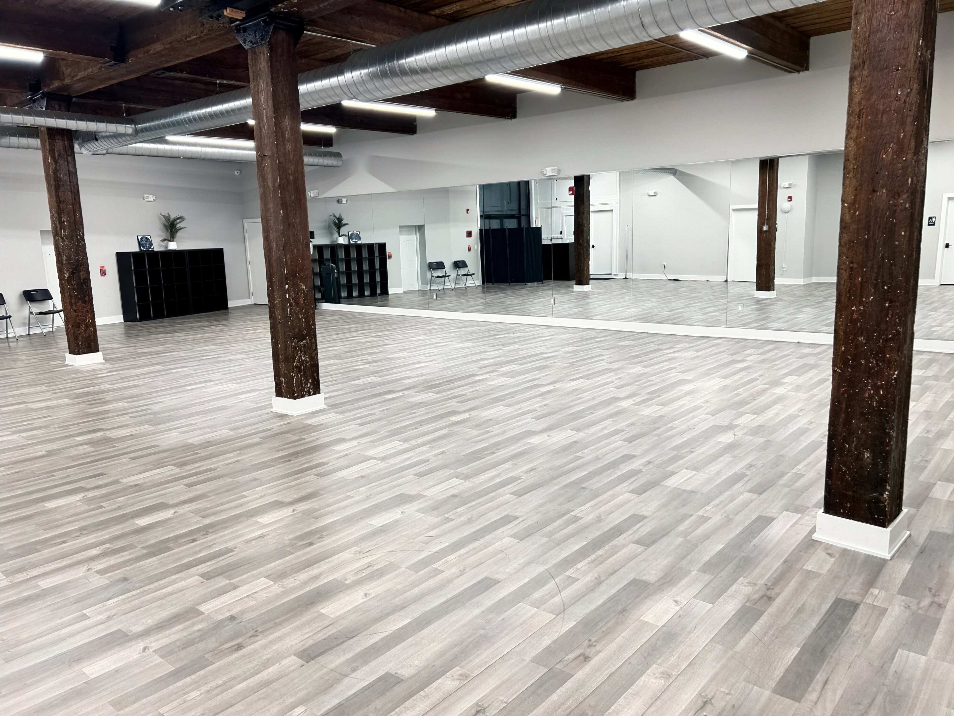 The image shows a spacious, empty dance studio with wooden floors, exposed beams, and a large mirror on one wall.