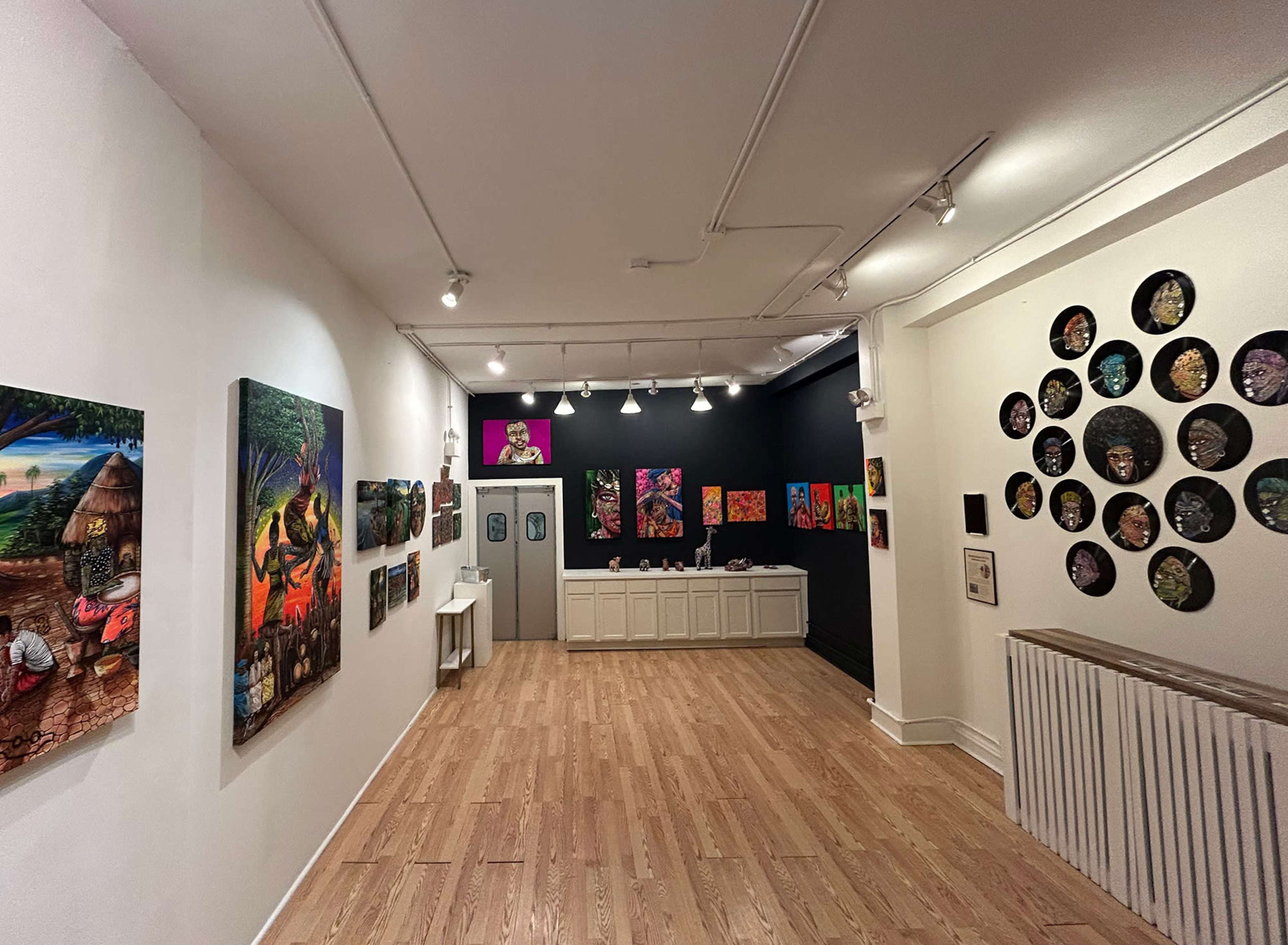 The image shows an art gallery with colorful paintings displayed on white and black walls, wooden flooring, and a reception area in the background.