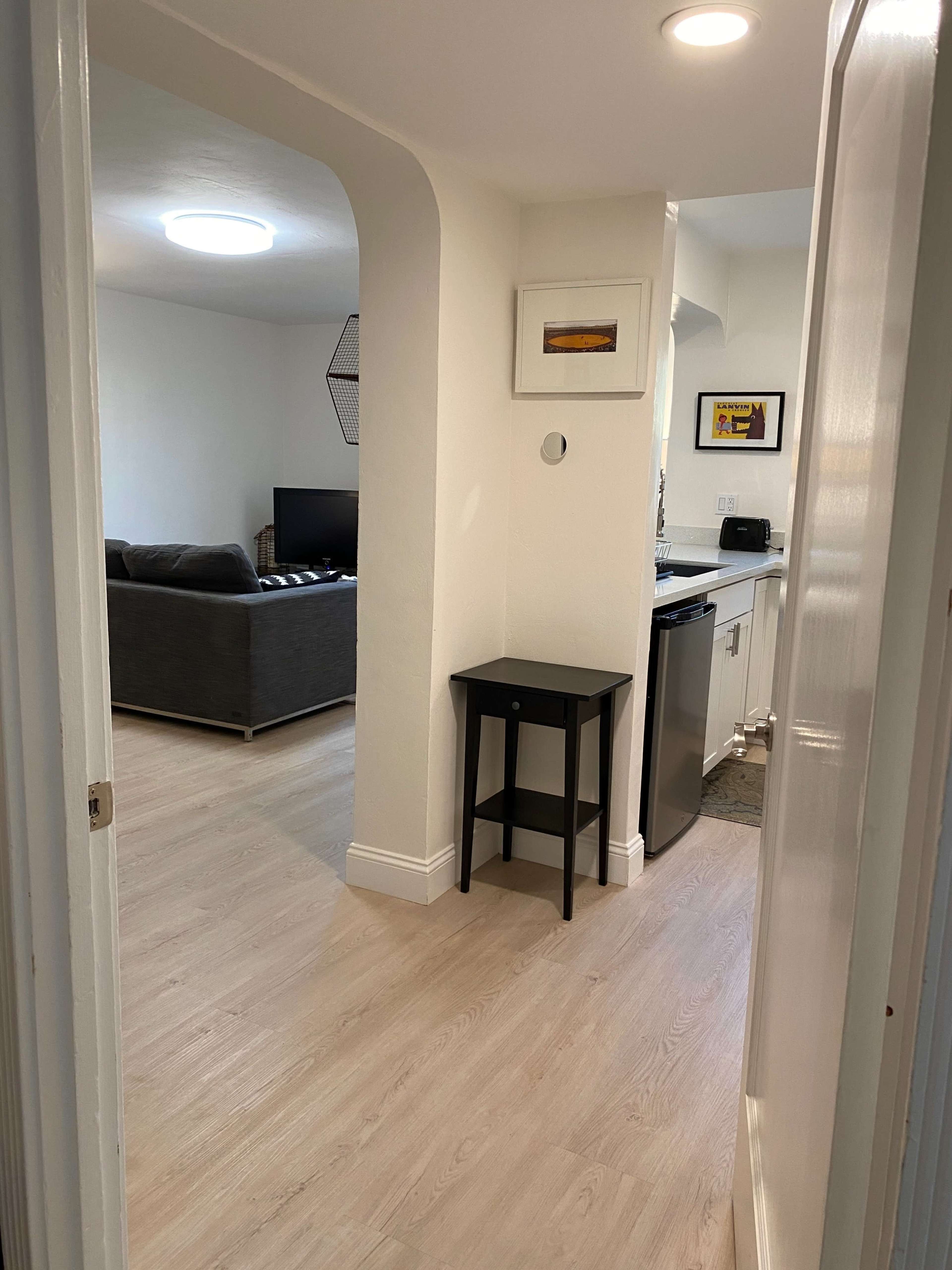 A narrow hallway leading to a living room with a gray sofa and a television, along with a kitchen area that includes a black table and stainless-steel appliances.