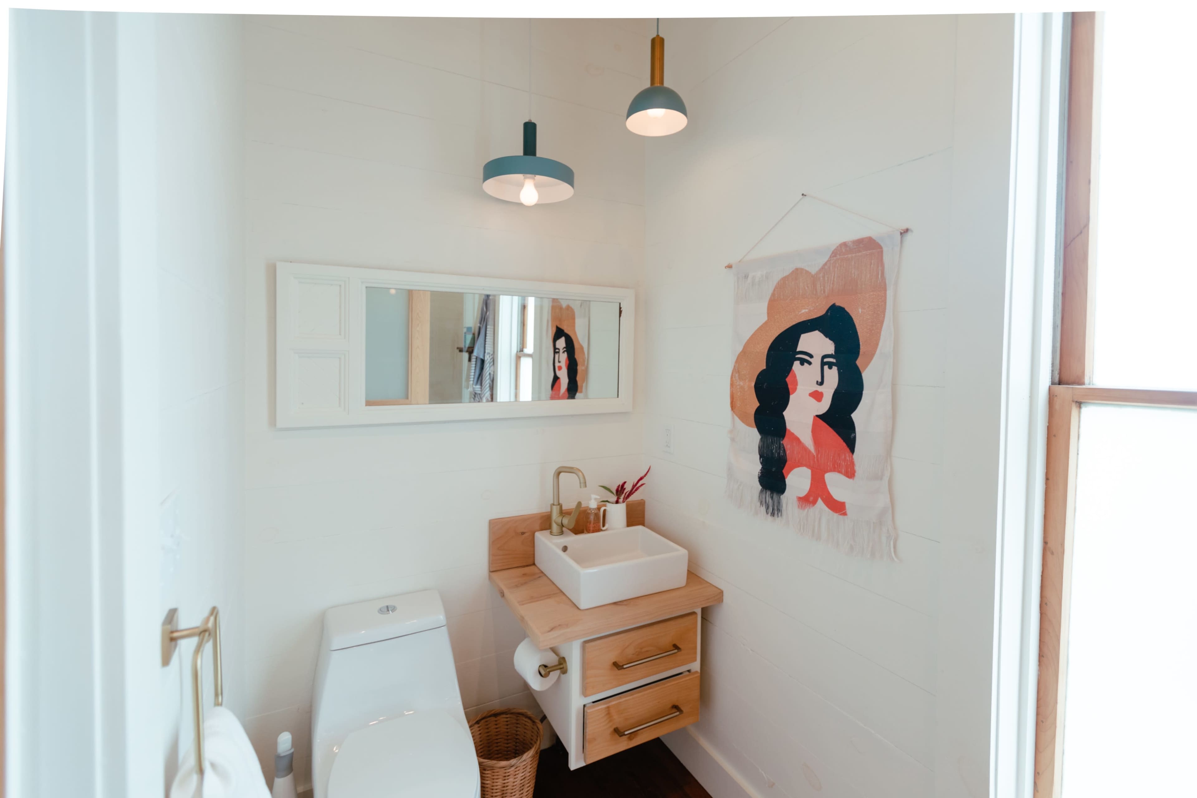 The bathroom features a wall-mounted sink with a wooden countertop, a mirror, artwork of a woman, and a toilet.